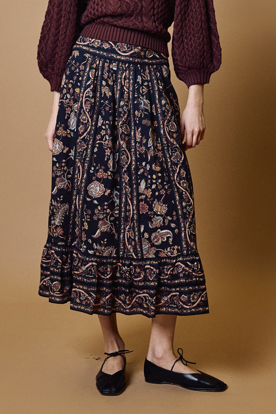 Wearing the Cleobella Morgan Midi Skirt in BOIS PRINT BOIS, a person stands against a brown background, paired with a maroon textured sweater and black ballet flats.