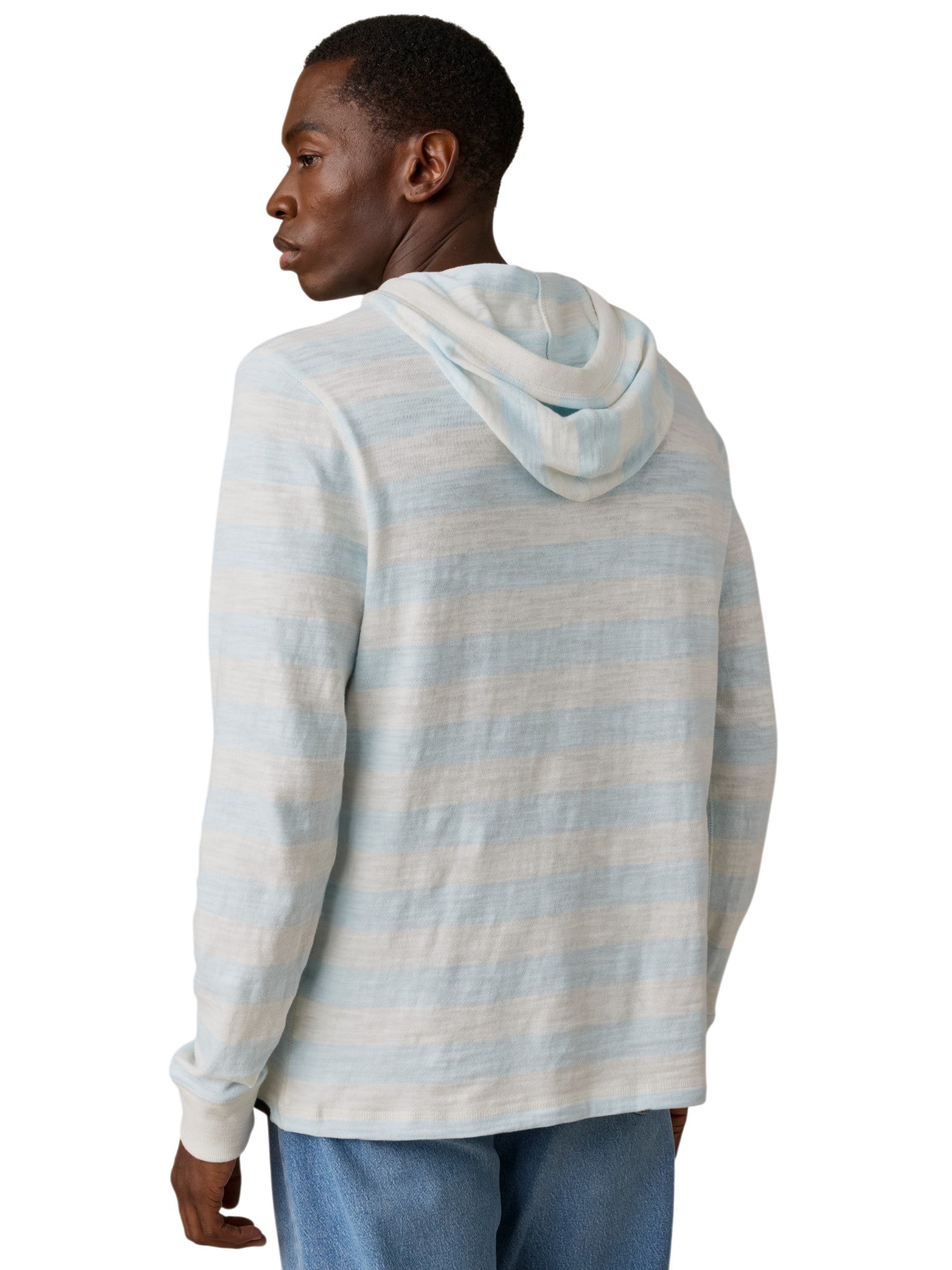 A man faces away, glancing over his shoulder. He wears the Faherty Sunwashed Slub Hoodie in light blue and white stripes with light blue jeans, set against a plain white background.