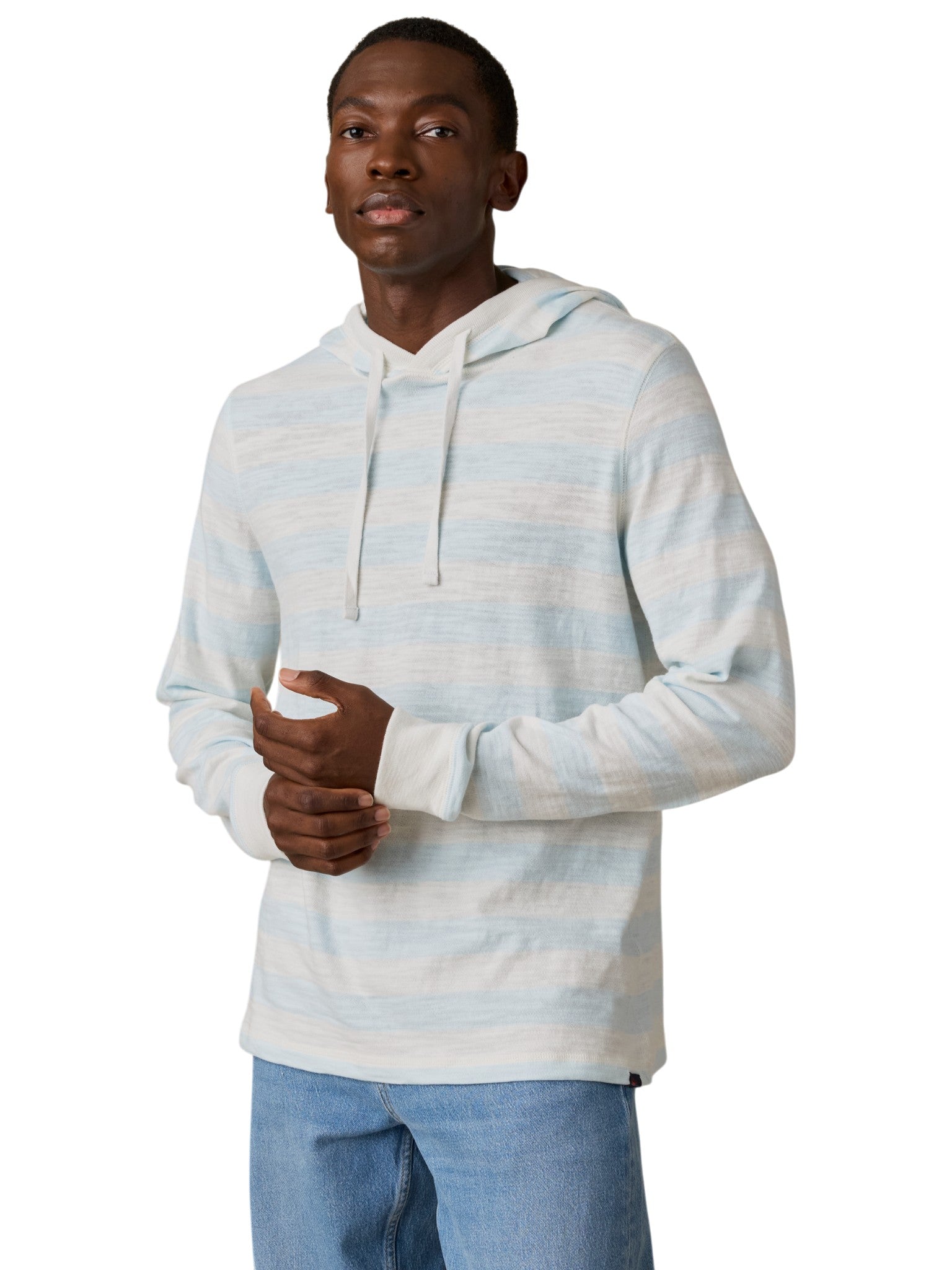 A man wears the Faherty Sunwashed Slub Hoodie, a relaxed-fit style in light blue and white stripes, paired with blue jeans. He stands against a plain white background, looking slightly to the side with a neutral expression.