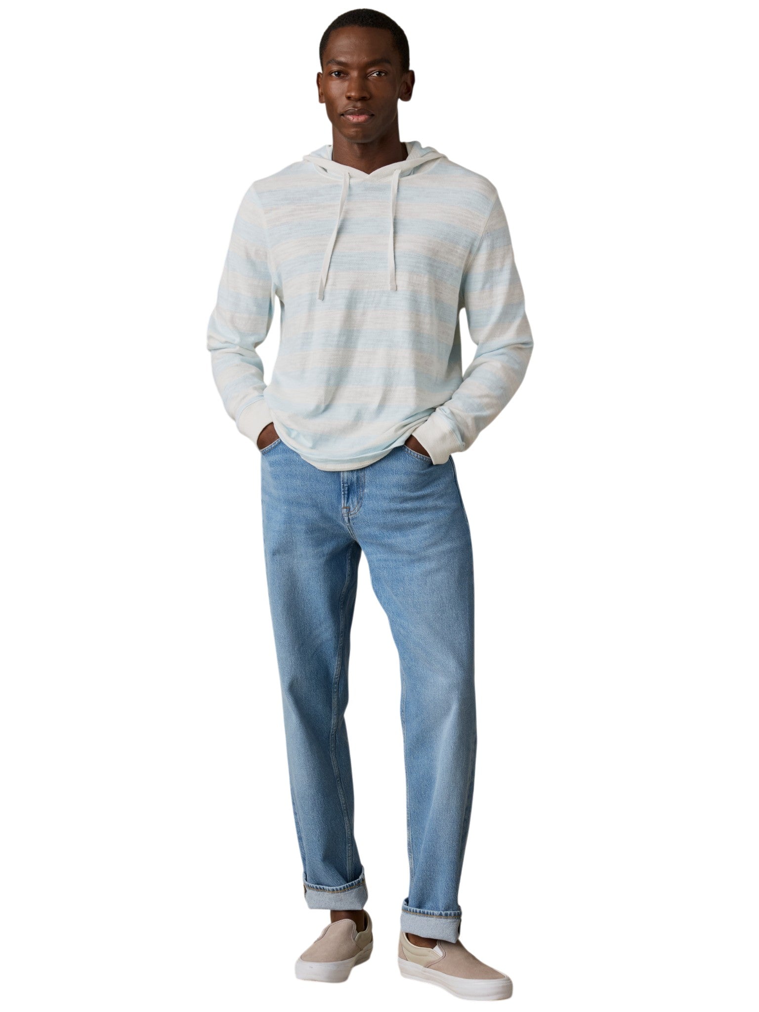 A man stands facing forward with his hands in his pockets, wearing the Faherty Sunwashed Slub Hoodie in a relaxed popover style, paired with light blue jeans and beige slip-on shoes. He has a neutral expression on his face.