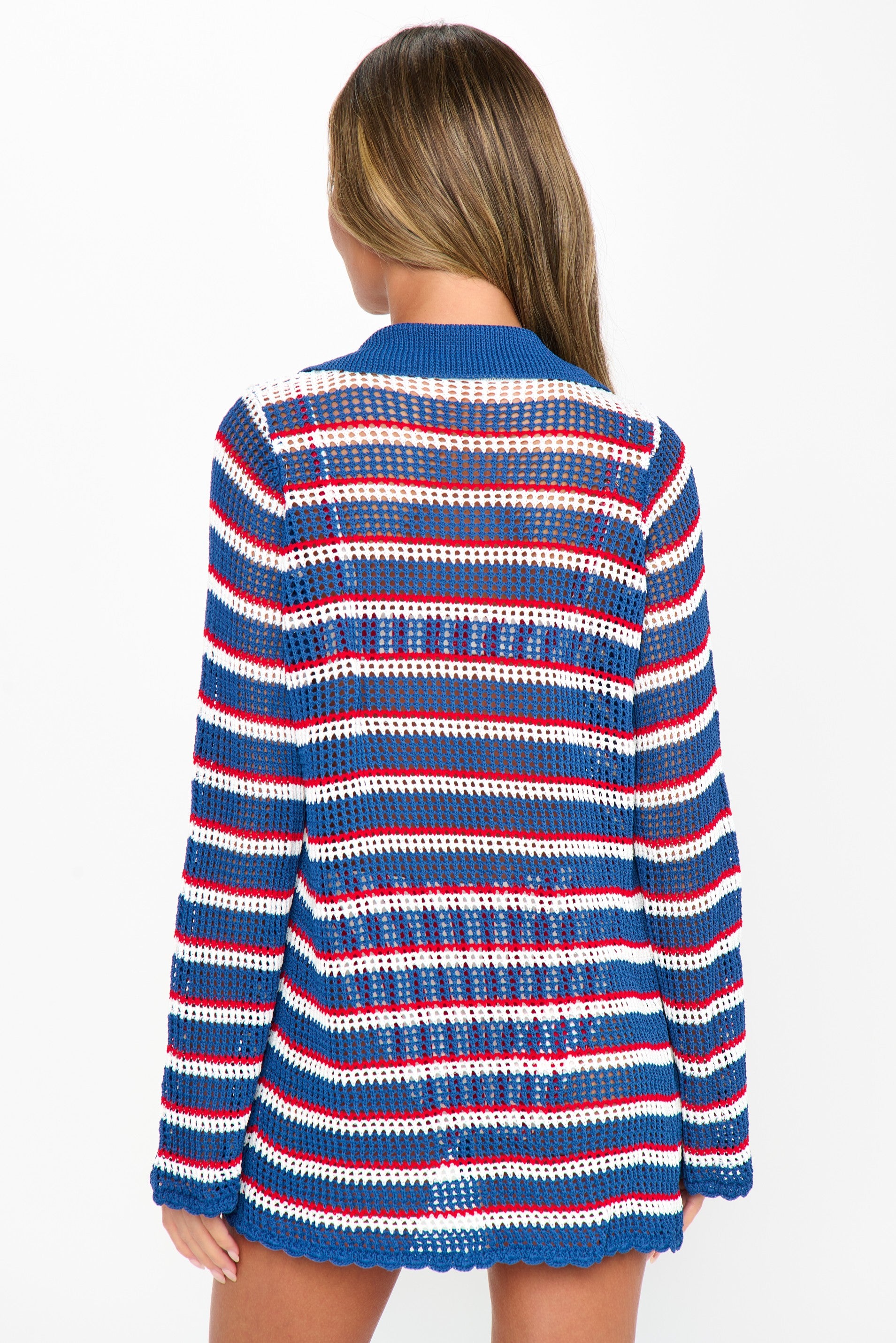 A person with long, light brown hair stands facing away, wearing the Show Me Your Mumu Button Up Coverup—a long-sleeve, open-knit beach sweater with horizontal red, white, and blue stripes—slightly sheer against a plain white background.