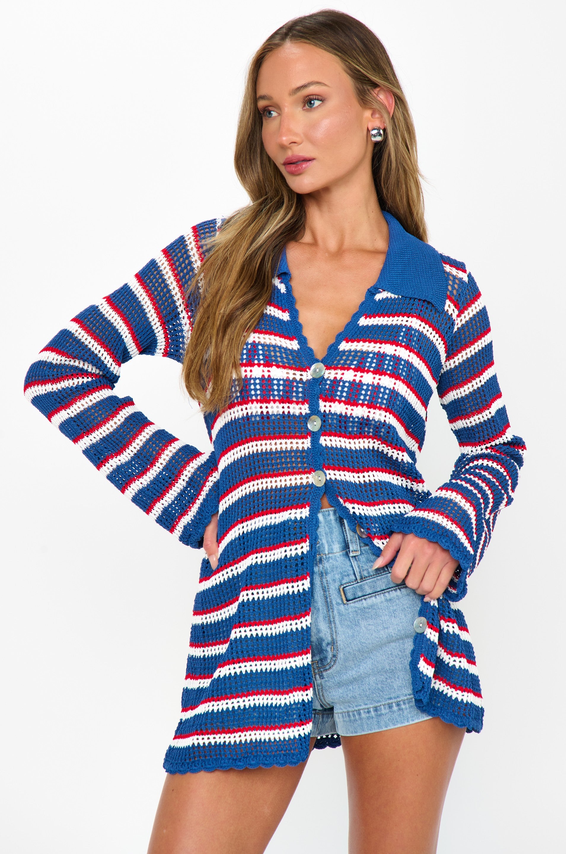 A woman models the Show Me Your Mumu Button Up Coverup—a long-sleeve, striped crochet shirt in blue, white, and red—over a blue tank and high-waisted denim shorts, styled against a crisp white background.