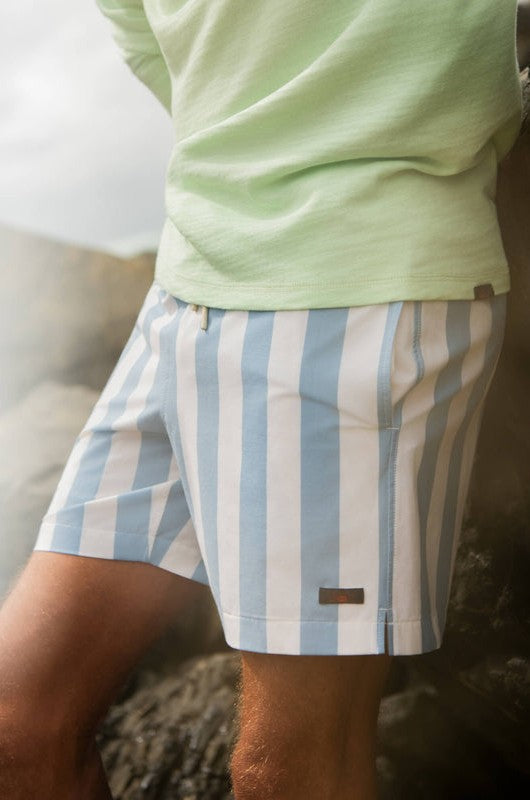 A person wears Faherty's All Day Trunk 6 In swim trunks—light blue and white vertical stripes with an elastic waistband—paired with a light green shirt, standing outdoors near rocks (only lower torso and upper legs shown).