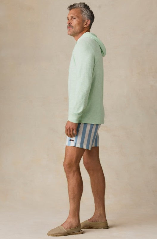 A man in profile wears a light green hoodie, Faherty All Day Trunk 6 In swim trunks in blue and white stripes, and beige slip-on shoes against a neutral background.