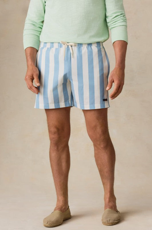A man stands on a neutral background wearing Faherty's All Day Trunk 6 In in BEACH BREEZE STRIPE, featuring blue-and-white vertical stripes and an elastic waistband. He pairs them with a light green long-sleeve shirt and beige slip-on shoes. Face not visible.