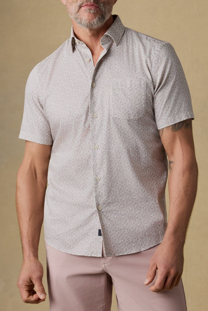A man with a gray beard wears the Faherty Short Sleeve Movement Monterrey Shirt and light pink pants, standing against a beige background.