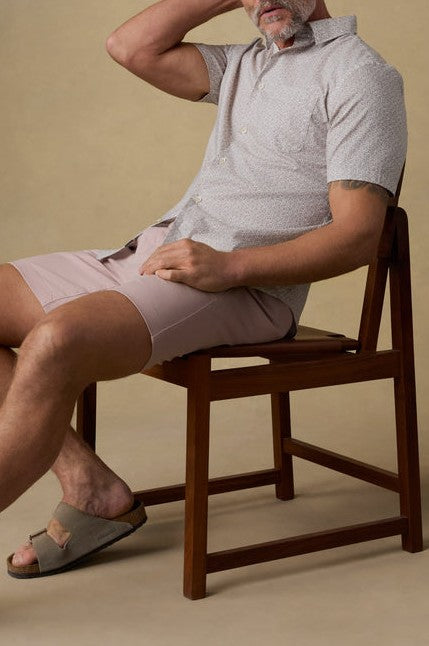A man with light skin and a gray beard sits on a wooden chair against a plain beige background, wearing the Faherty Short Sleeve Movement Monterrey Shirt, light pink shorts, and beige sandals.