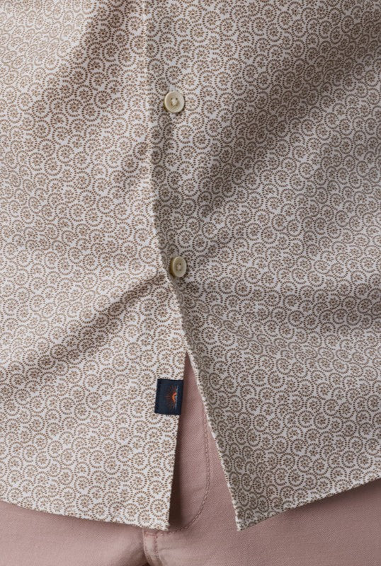 Close-up of the Faherty Short Sleeve Movement Monterrey Shirt, featuring a relaxed fit, spread collar, beige pattern, cream buttons, and a navy tag near the hem. The open bottom reveals pink pants underneath.