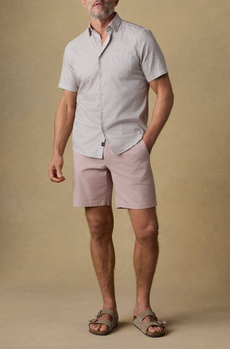 A man stands against a beige background wearing Faherty’s Short Sleeve Movement Monterrey Shirt, paired with light pink shorts and taupe sandals. His left hand hangs relaxed by his side while his right is slightly clenched.