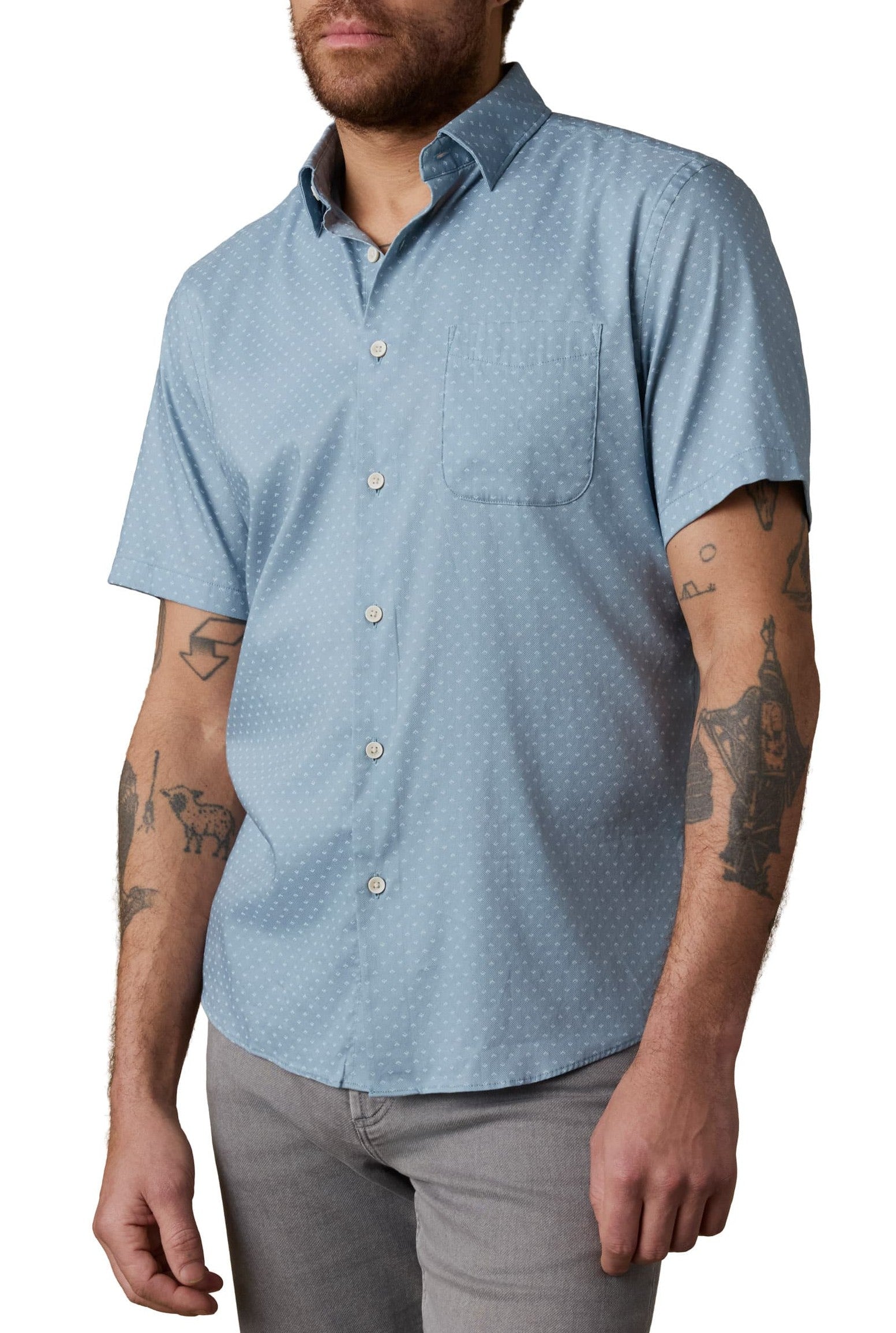 A man with arm tattoos wears the Faherty Short Sleeve Movement Monterrey Shirt, a relaxed fit light blue shirt with a subtle dotted pattern, spread collar, and chest pocket, paired with light gray pants.