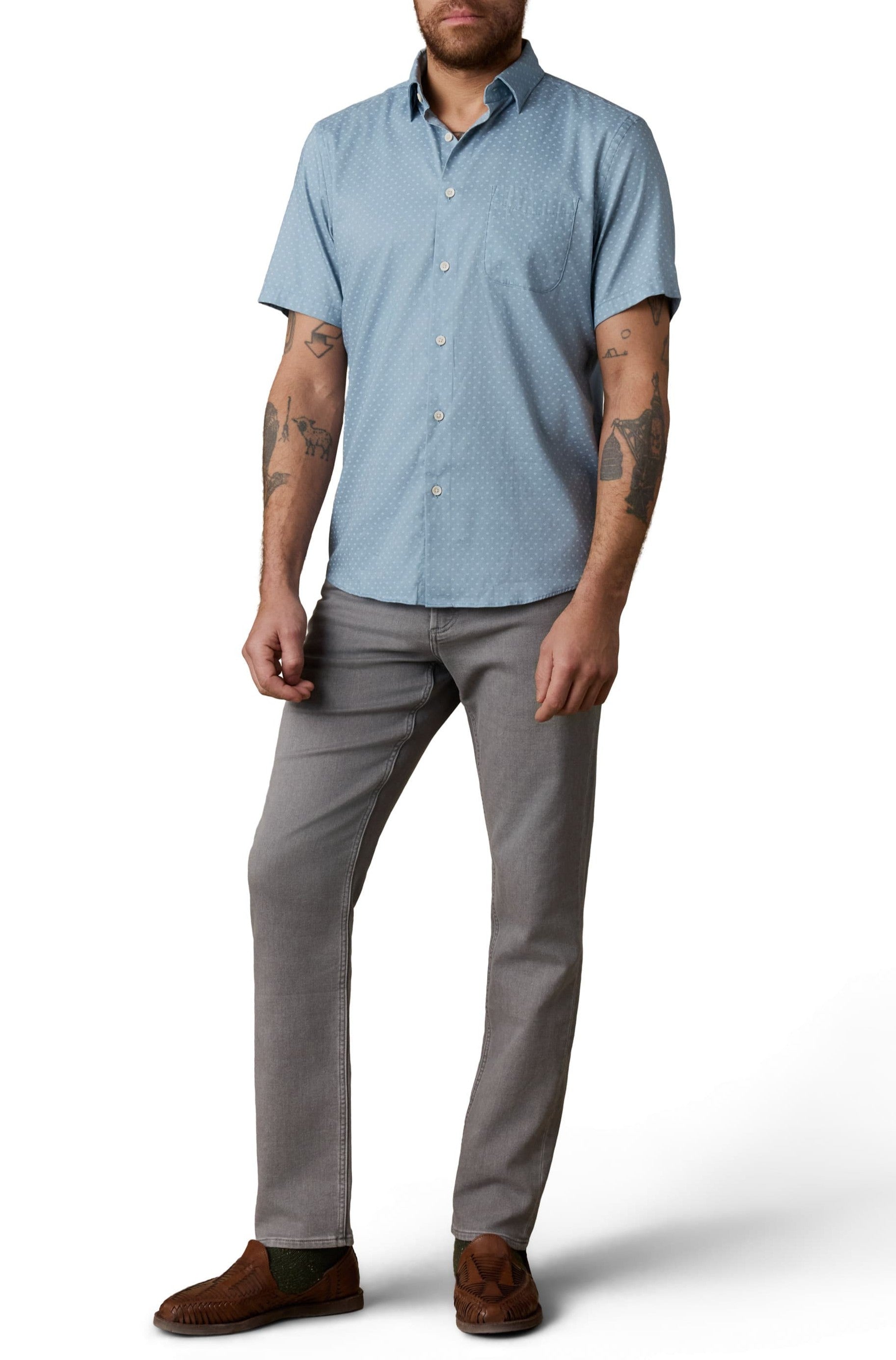 A man with tattooed arms wears a Faherty Short Sleeve Movement Monterrey Shirt in light blue, paired with gray pants and brown woven shoes, standing against a white background.