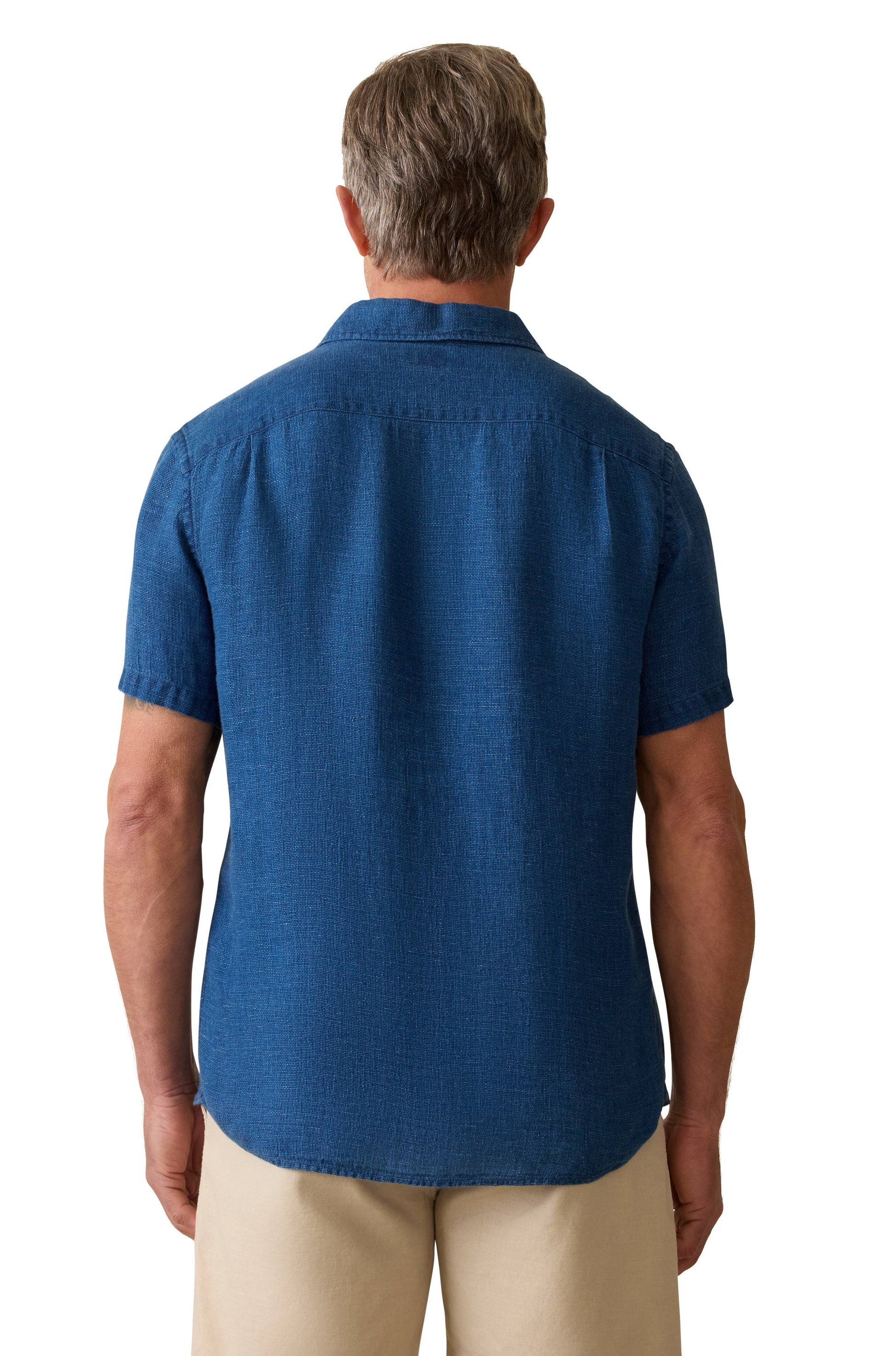 A man with short gray hair is seen from behind, wearing the Faherty Short Sleeve Palma Linen Shirt in blue with coconut buttons and beige pants, posed against a white background.