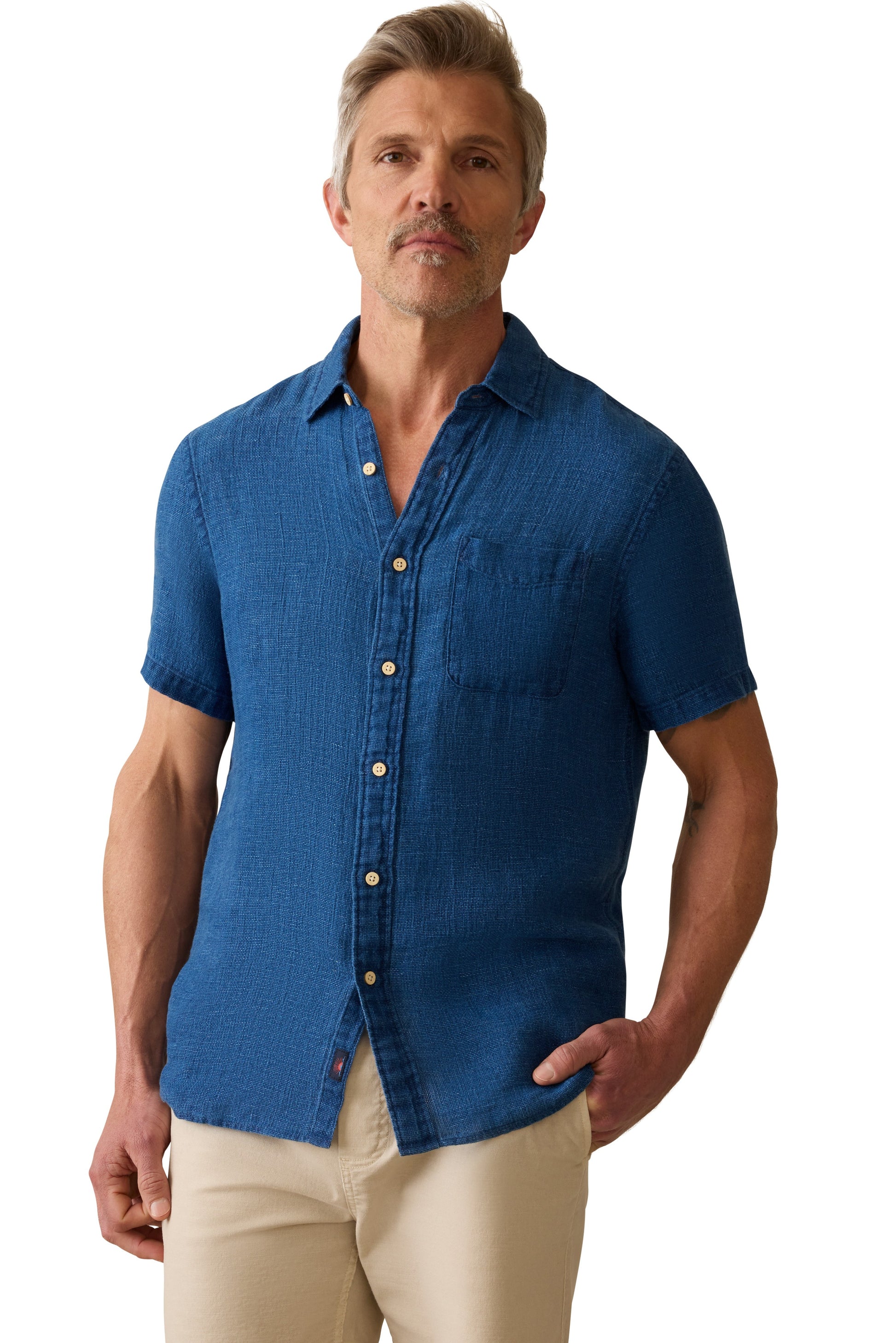 A man with short, light brown hair and a mustache wears the Faherty Short Sleeve Palma Linen Shirt in blue, featuring coconut buttons and a chest pocket. He pairs it with beige pants and stands against a white background with one hand in his pocket.