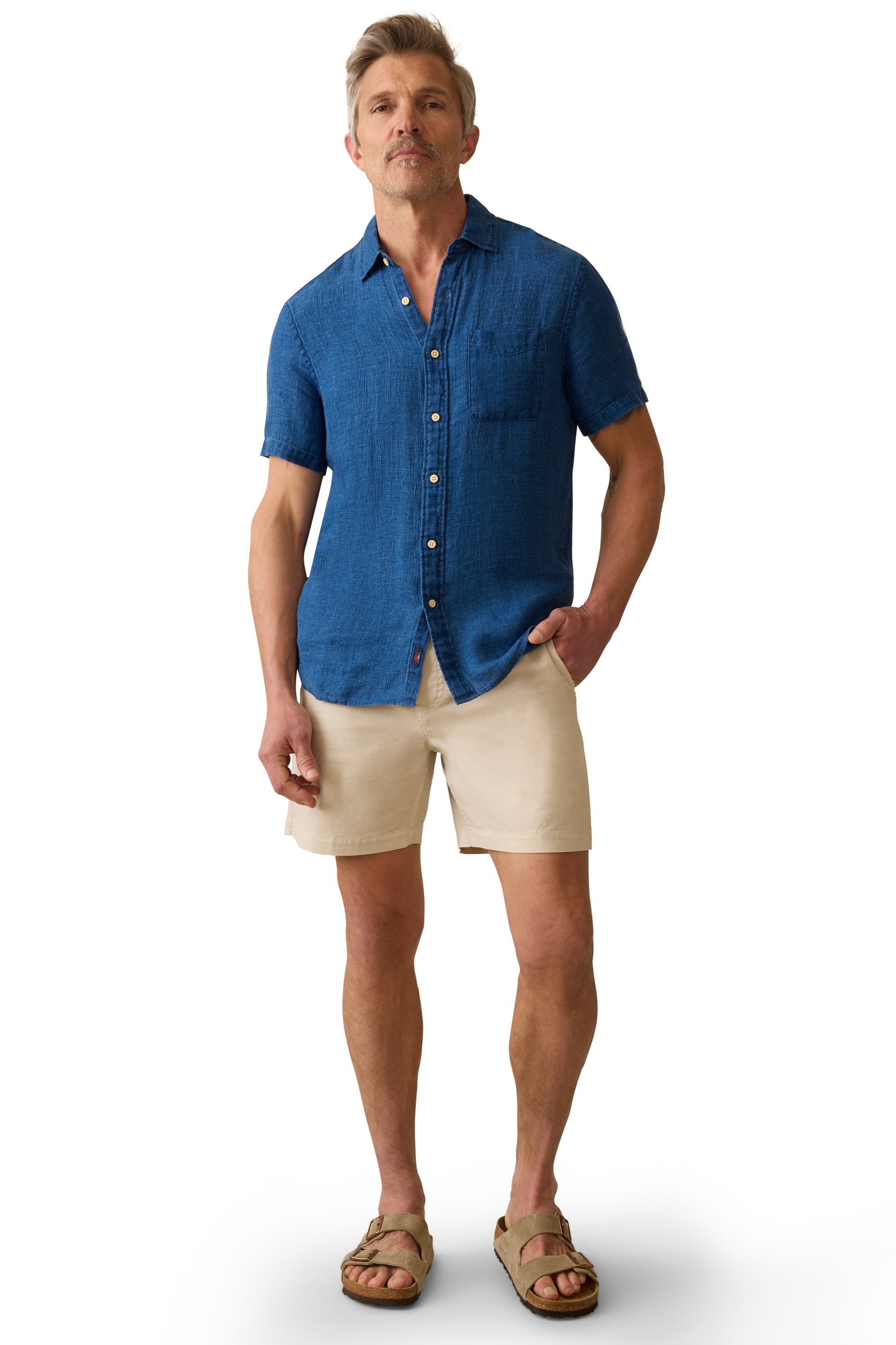 A man with a neutral expression stands against a white background, wearing the Faherty Short Sleeve Palma Linen Shirt in blue with coconut buttons, paired with beige shorts and sandals. One hand is in his pocket.