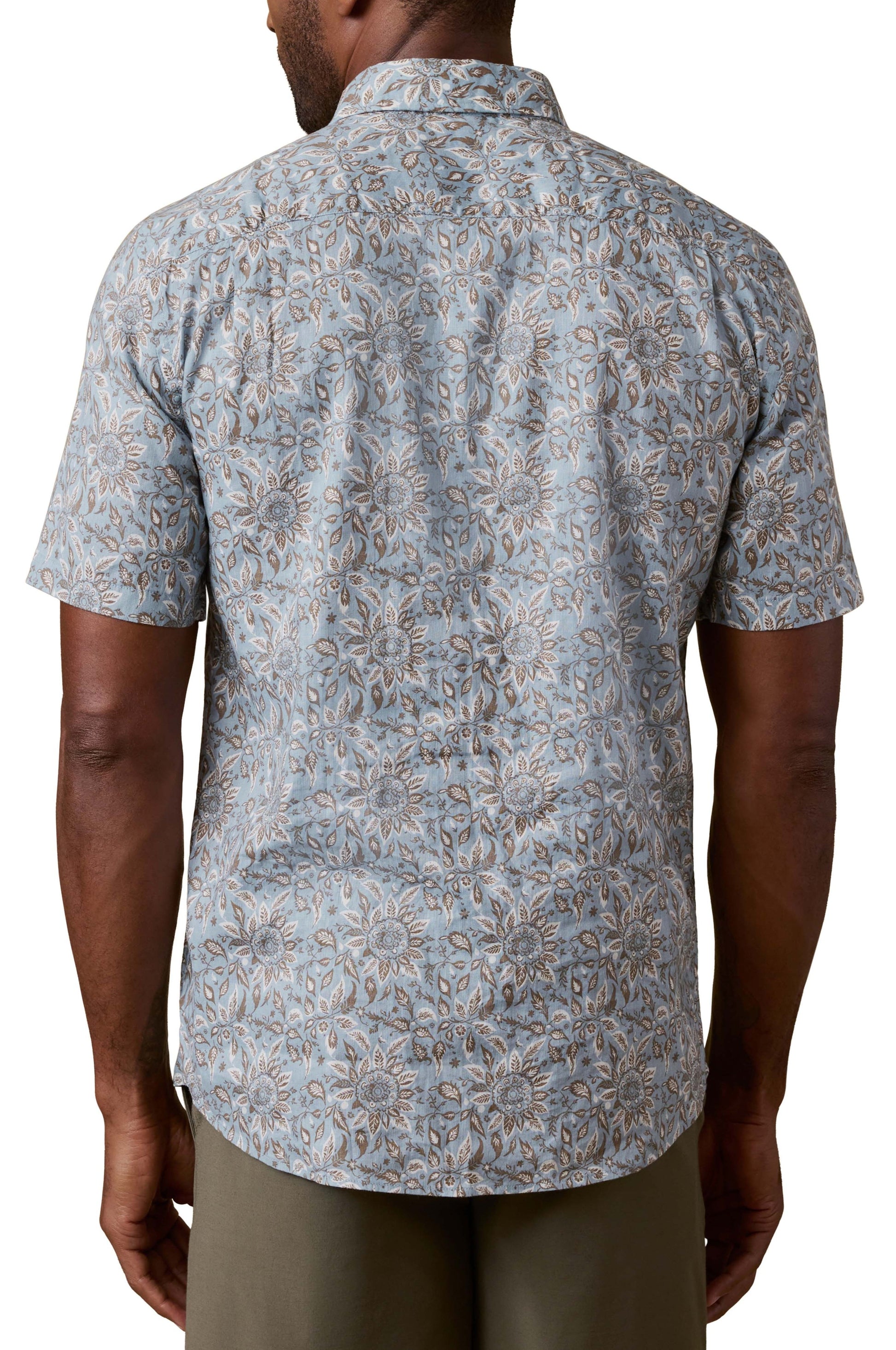 A man is shown from the back wearing the Faherty Short Sleeve Breeze Shirt, featuring a blue and beige floral pattern, relaxed fit, collar, and short sleeves. He pairs it with olive green pants for an easygoing vacation look.