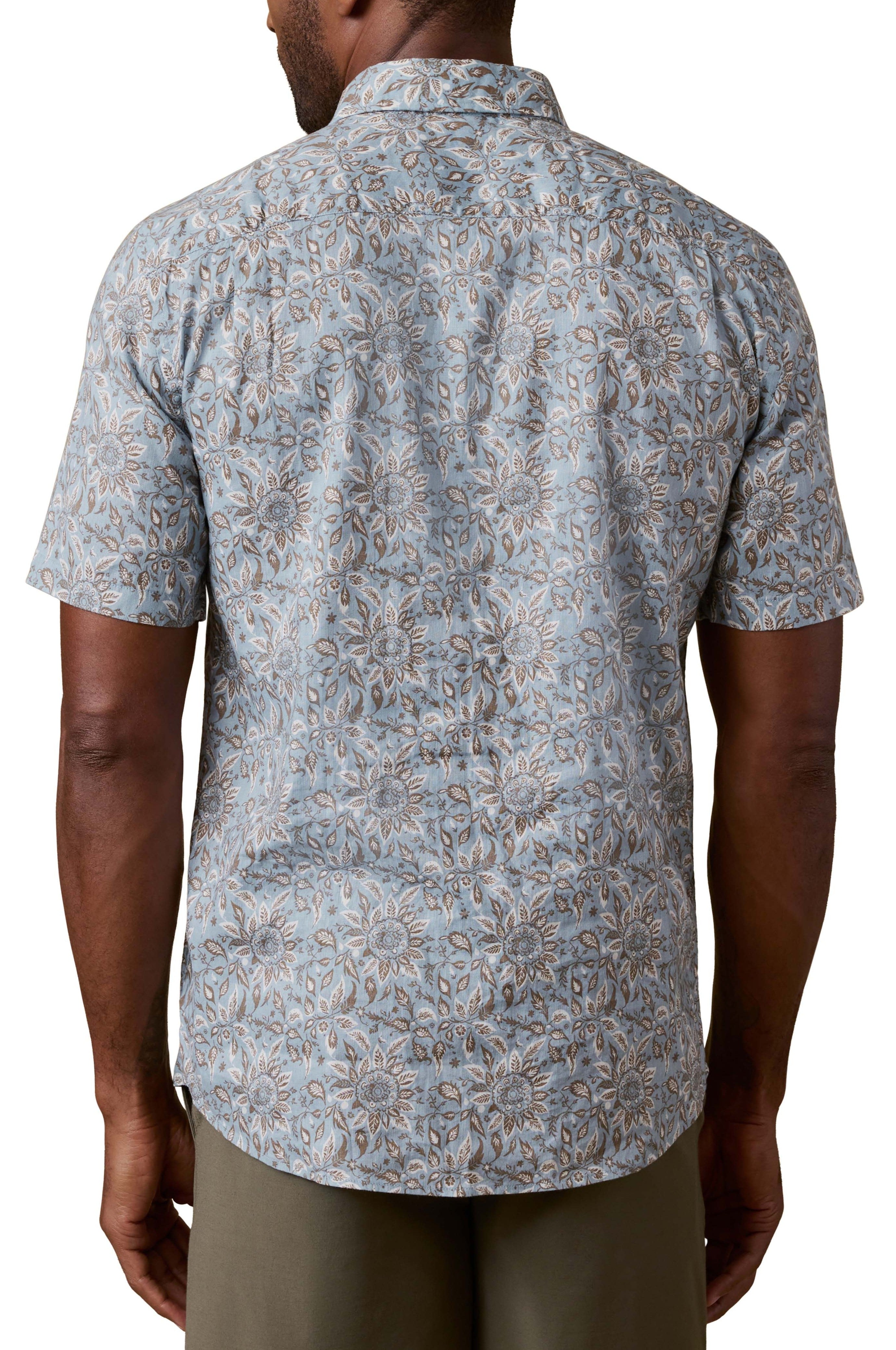 A man is shown from the back wearing the Faherty Short Sleeve Breeze Shirt, featuring a blue and beige floral pattern, relaxed fit, collar, and short sleeves. He pairs it with olive green pants for an easygoing vacation look.