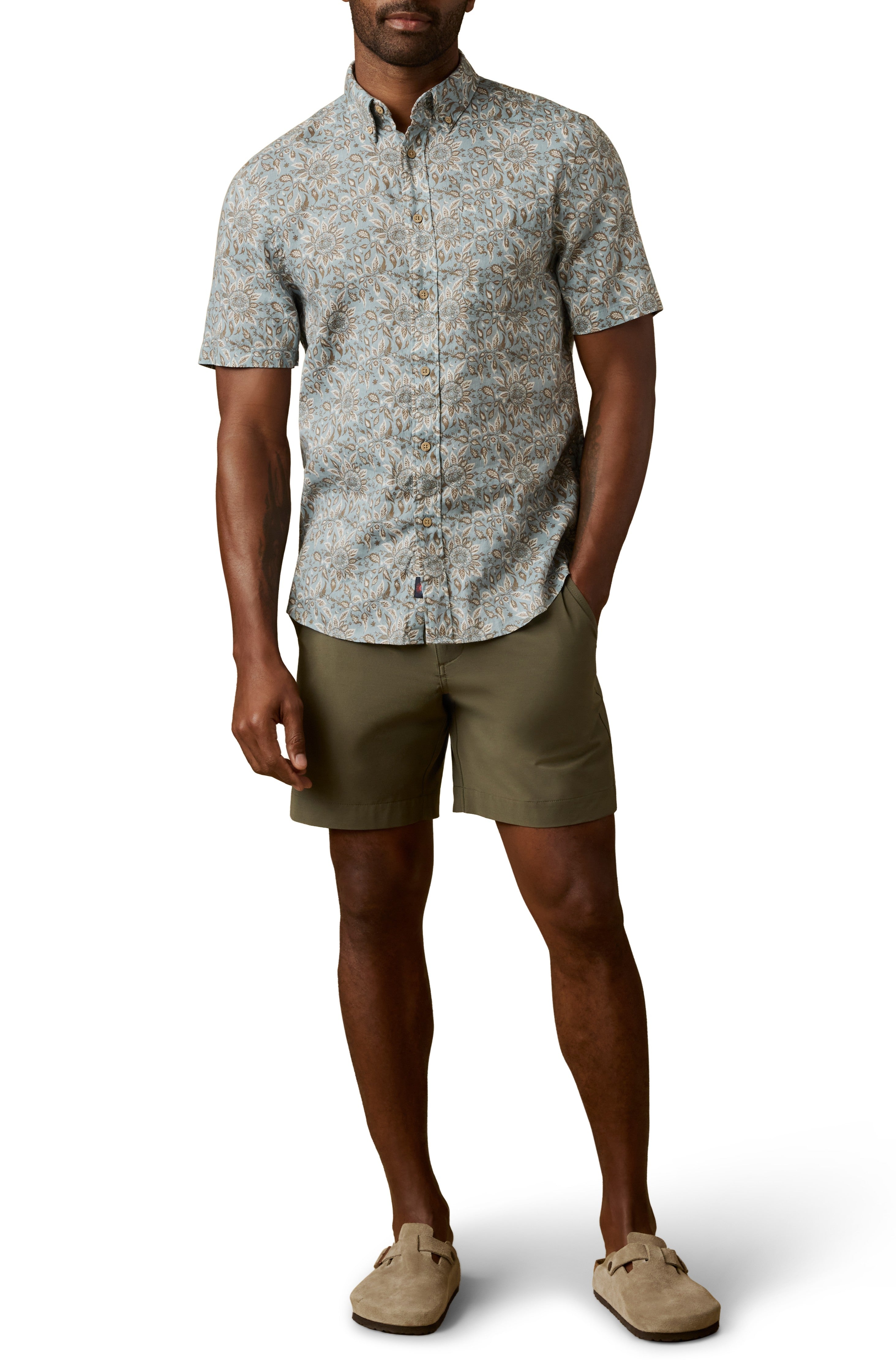 A man in a Faherty Short Sleeve Breeze Shirt, featuring a light blue pattern, pairs it with olive green shorts and beige slip-on shoes, standing with one hand in his pocket against a white background.