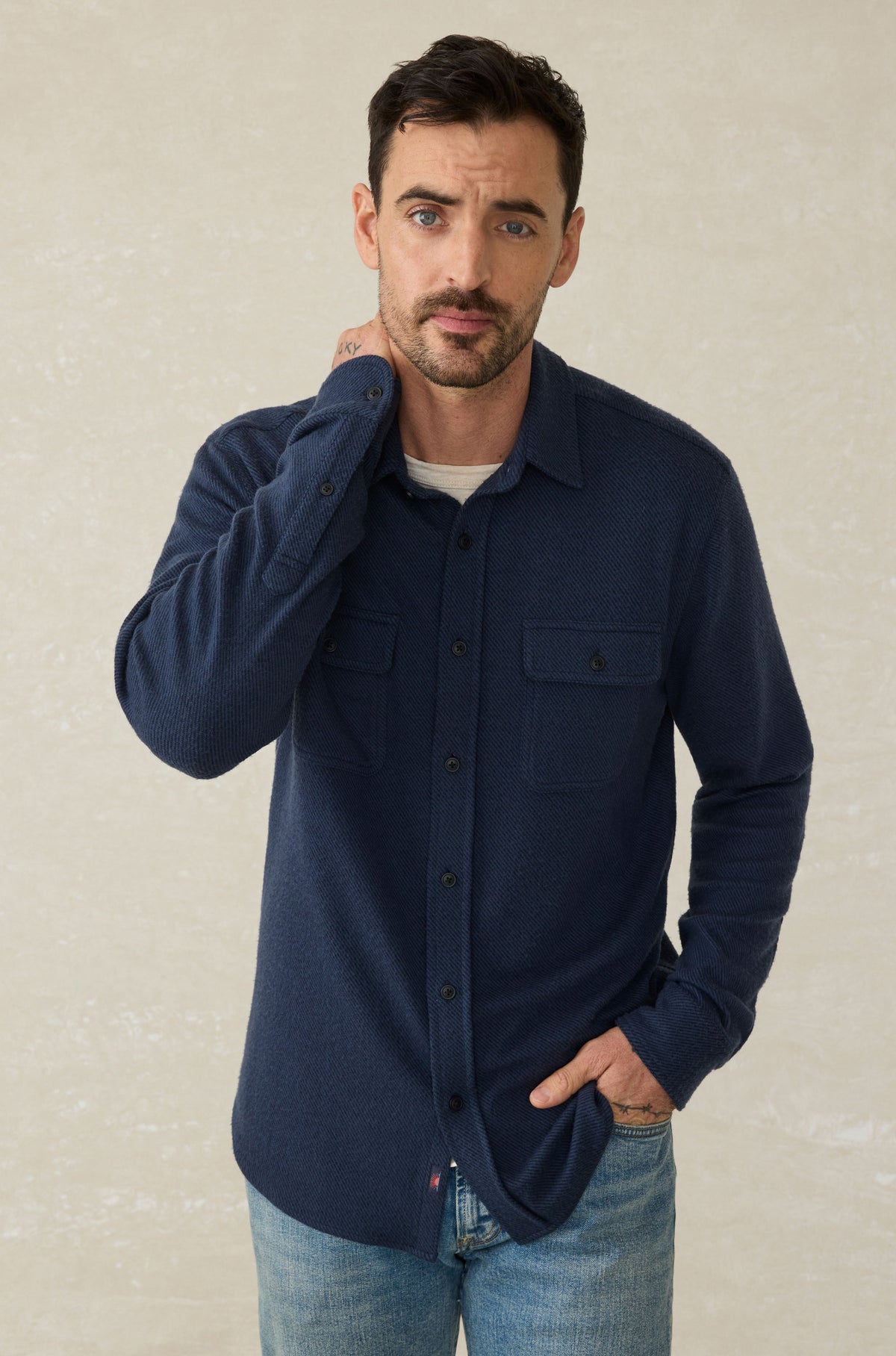 Man wearing navy blue textured button-up shirt with chest pockets over white undershirt and light blue jeans.