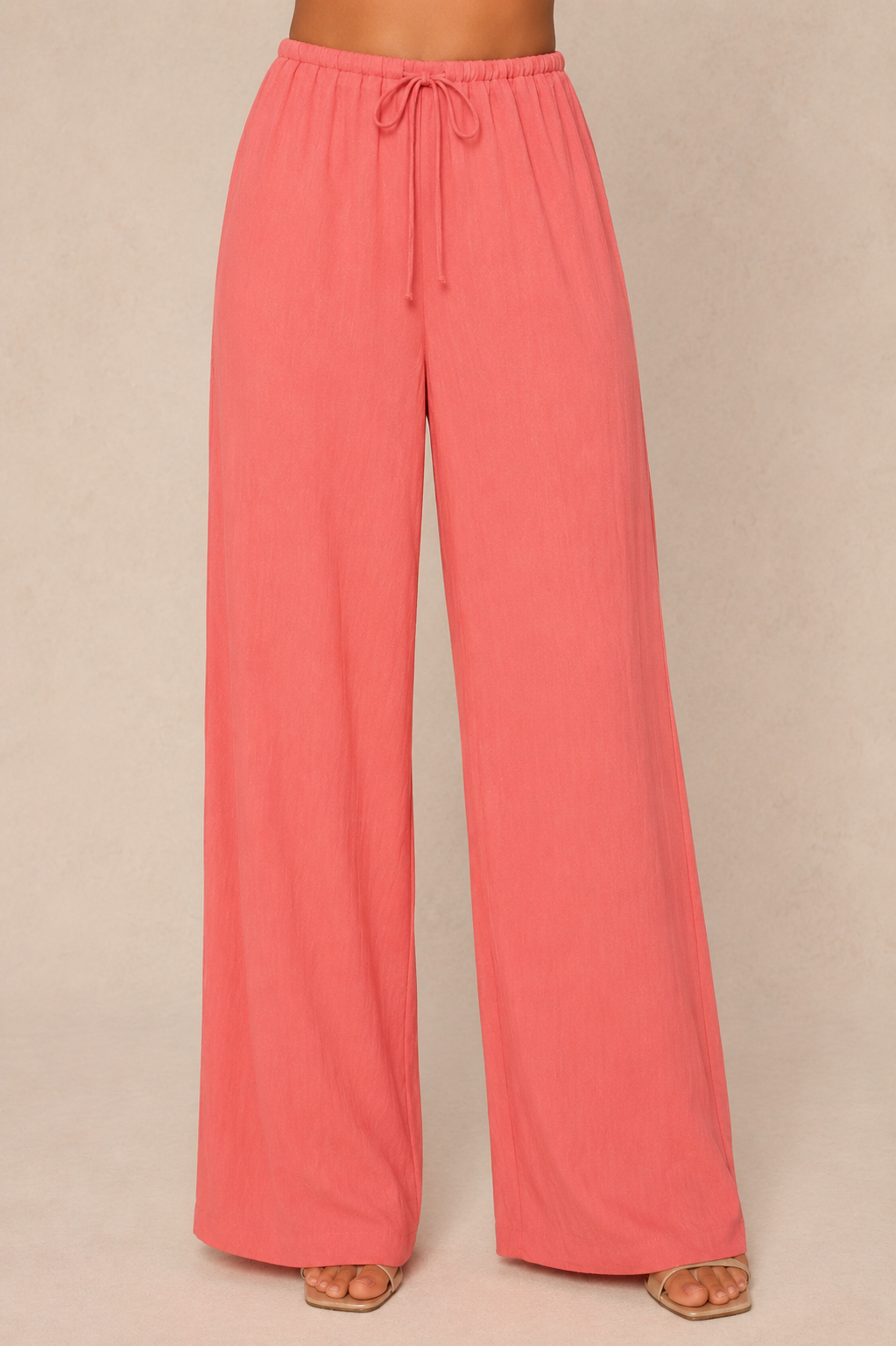 A person wears the Georgie Grayton Beach Pant in CORAL—a loose, high-waisted, wide leg style with an elastic drawstring waist—standing on a light beige background. The flowy pants reach the floor.