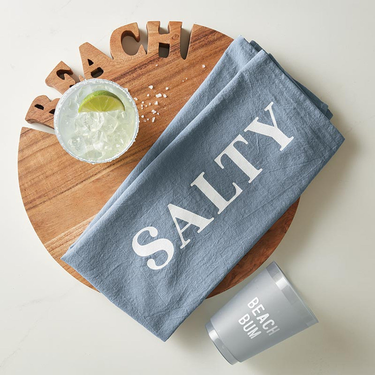 A gray cloth with SALTY printed on it is set on a round wooden board beside Santa Barbara Design Studio's Beach Bum Frosted Cups (8 Ct)—including a gray BEACH BUM cup, lime-garnished drinks, and wooden letters spelling BEACHY.