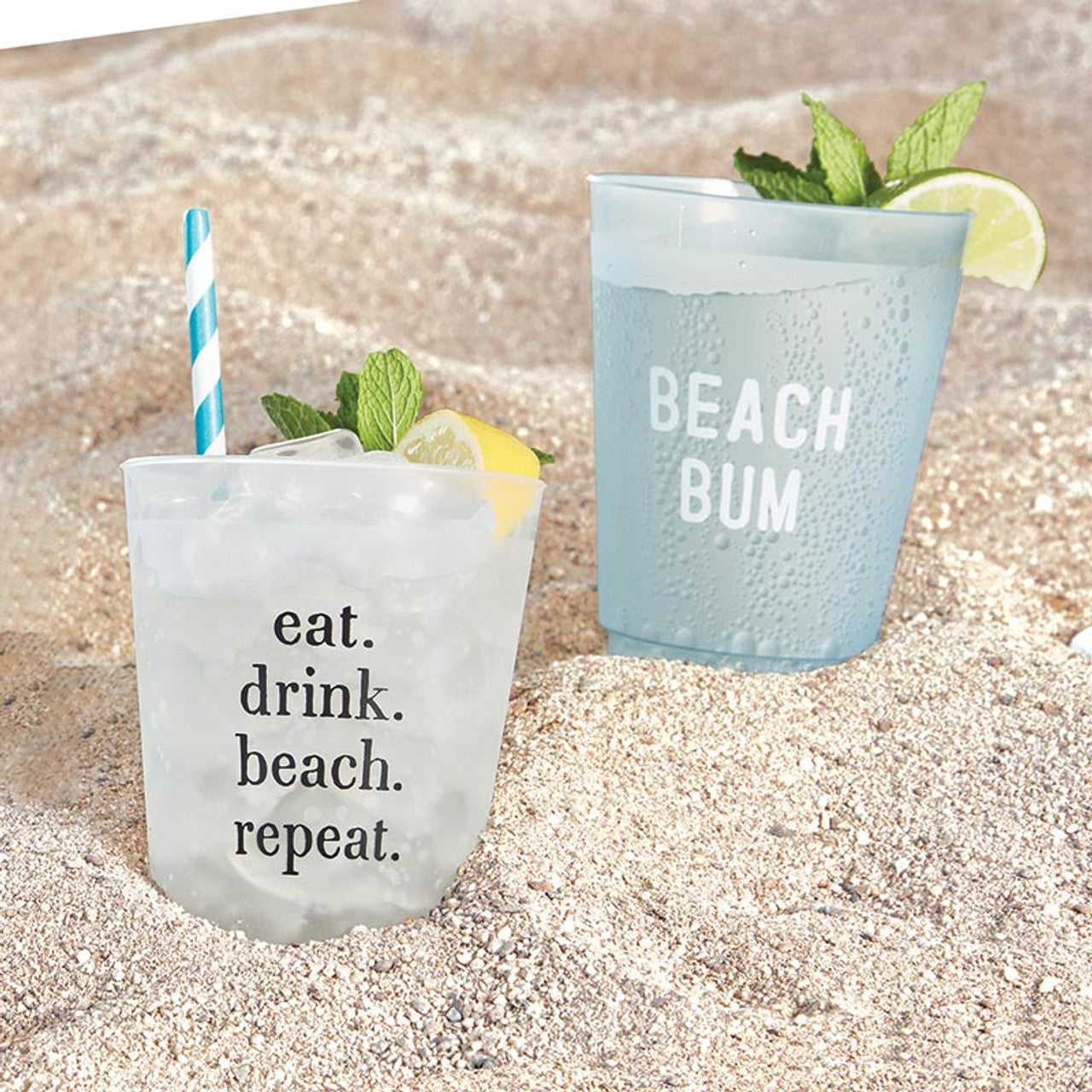 Two Santa Barbara Design Studio Beach Bum Frosted Cups with ice drinks are on a sandy beach, one reading "eat. drink. beach. repeat." with a blue striped straw and lemon; the other says "BEACH BUM" with mint and lime garnish. Set of 8 cups.