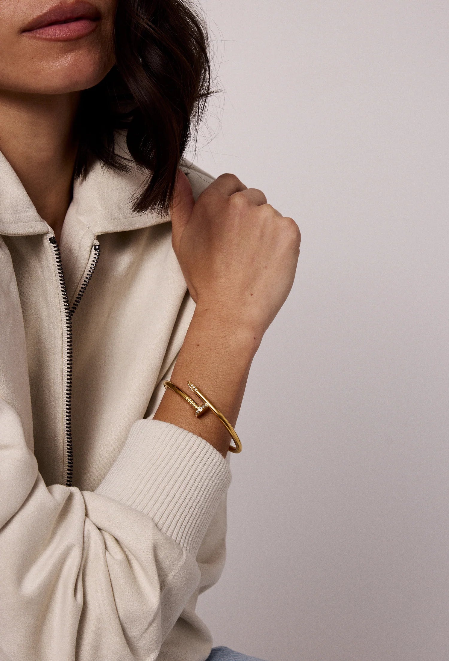 A woman in a cream zip-up hoodie rests her left arm on her chest, highlighting the 4mm thick, 14kt gold plated Nailed It Bracelet by Electric Picks on her wrist. Her face is partly visible with shoulder-length dark hair.