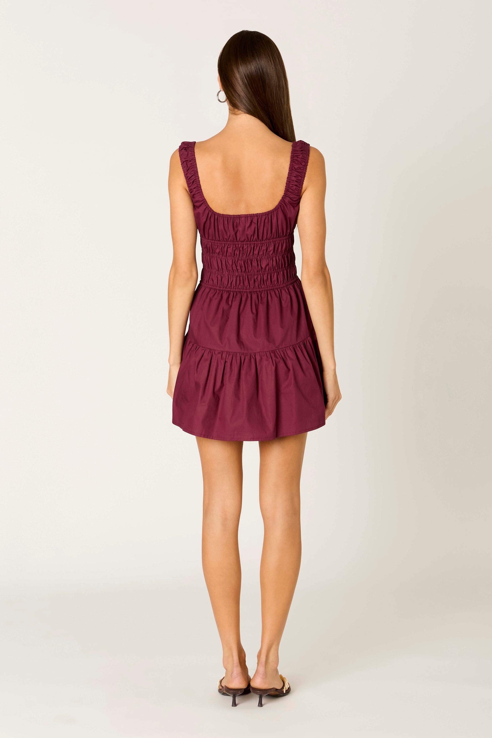 A woman with long brown hair is standing with her back to the camera against a plain light background, wearing the Nia Salt Mini Dress—a sleeveless, short maroon fit-and-flare dress with a smocked bodice—paired with heeled sandals.