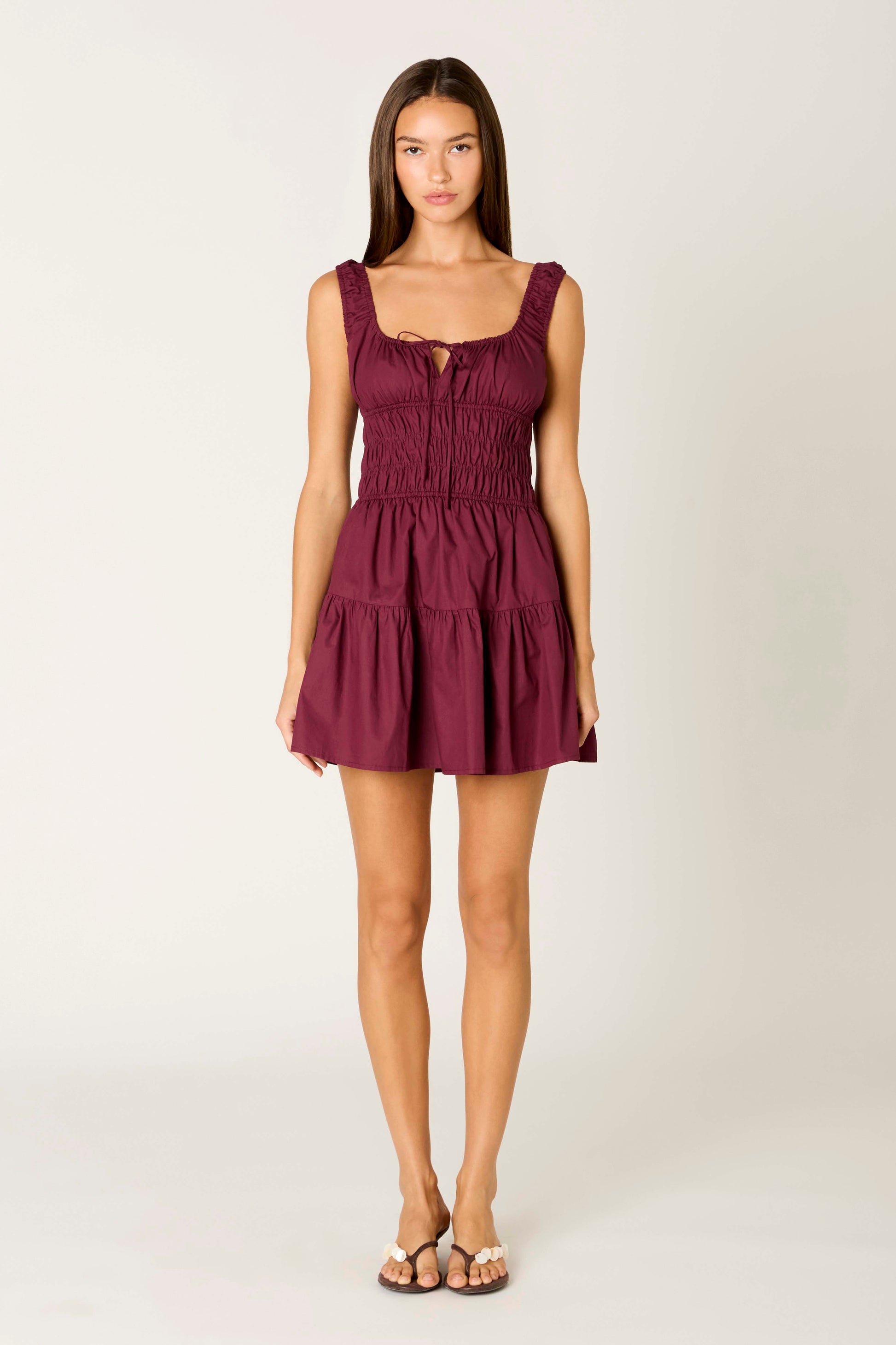 A woman stands facing forward against a plain light background, wearing the Salt Mini Dress by Nia in PLUM—a sleeveless, short fit-and-flare style featuring a fitted ruched bodice and ruffled straps—paired with flat sandals.