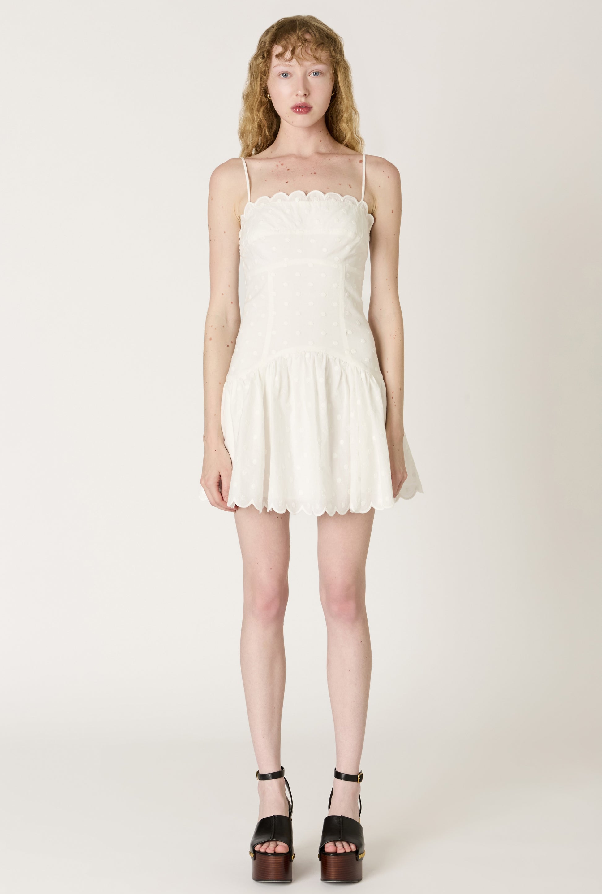 A light-skinned woman with wavy blonde hair wears the white Elvira Mini Dress by Nia, featuring scalloped edges, subtle polka dots, and a fit-and-flare silhouette, paired with black high-heeled sandals against a plain light background.