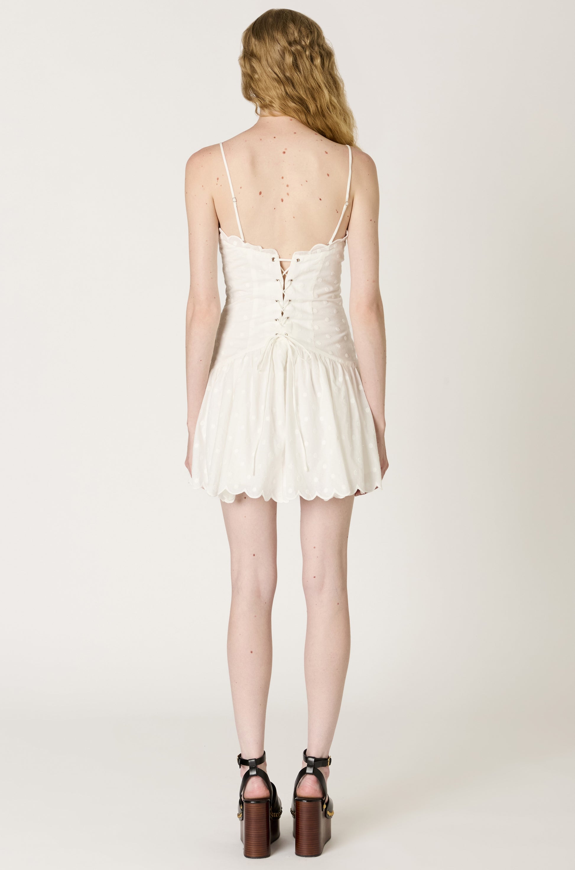 A woman with wavy, light brown hair is shown from the back in a Nia Elvira Mini Dress with thin straps and a lace-up corset back, paired with black platform ankle-strap heels. The background is plain white.