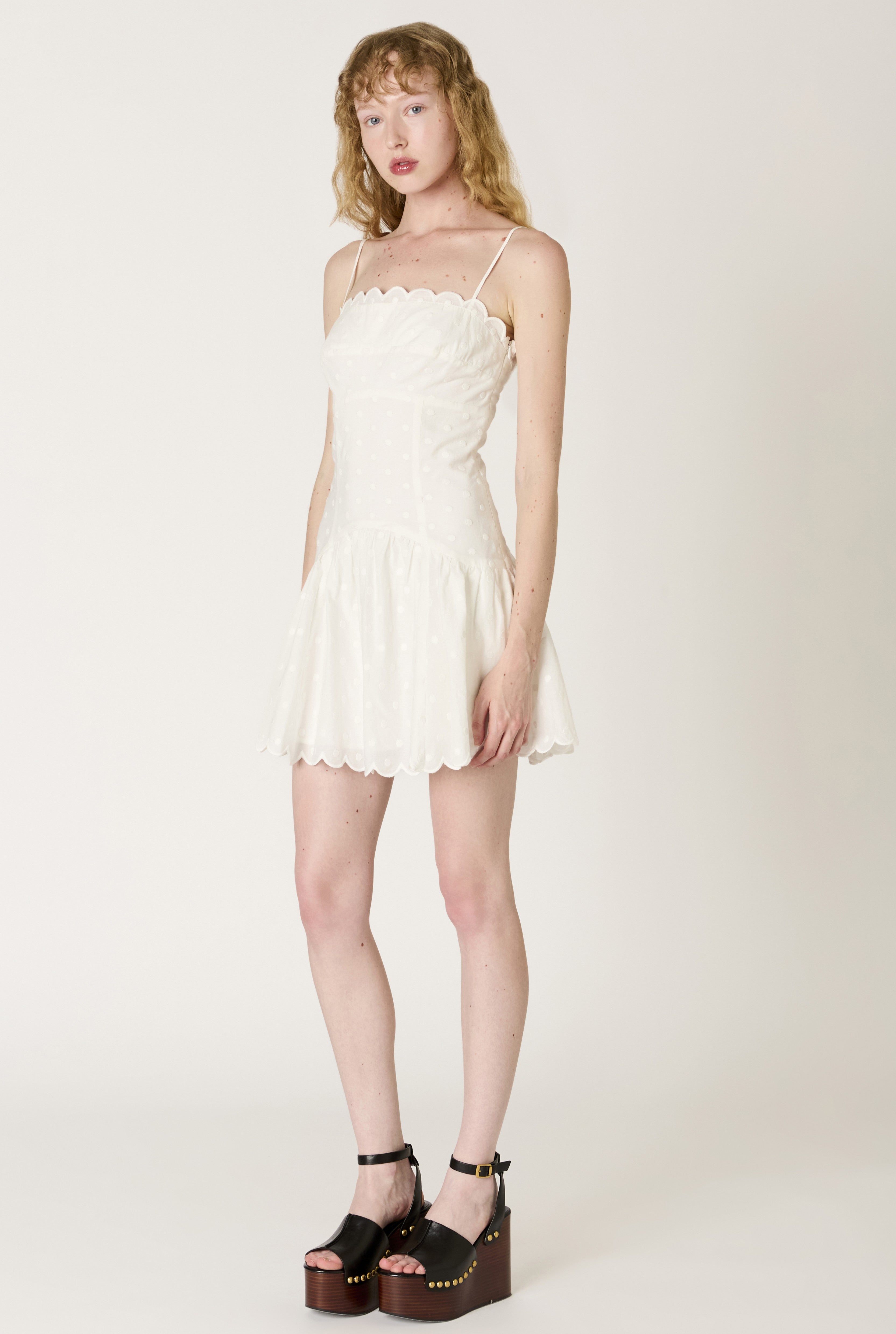 A woman wears the Elvira Mini Dress by Nia, a white fit-and-flare mini with thin straps, scalloped hem, and subtle polka dots, paired with black platform sandals. Her wavy light brown hair frames her look against a plain background.