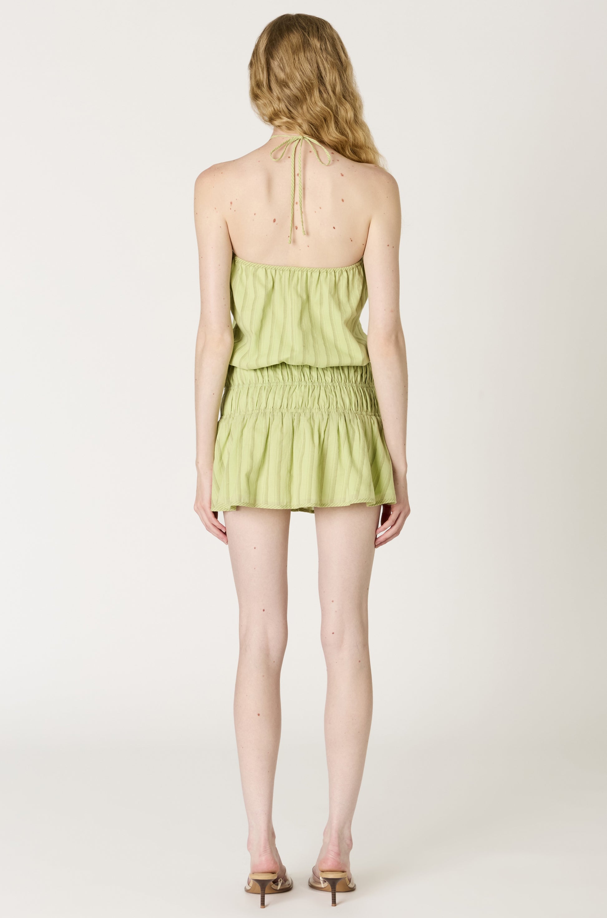A woman with wavy blonde hair faces away, wearing the Nia Mara Dress Coverup—a light green cotton mini dress with a halter neck and open back—paired with beige heeled sandals against a plain white background.