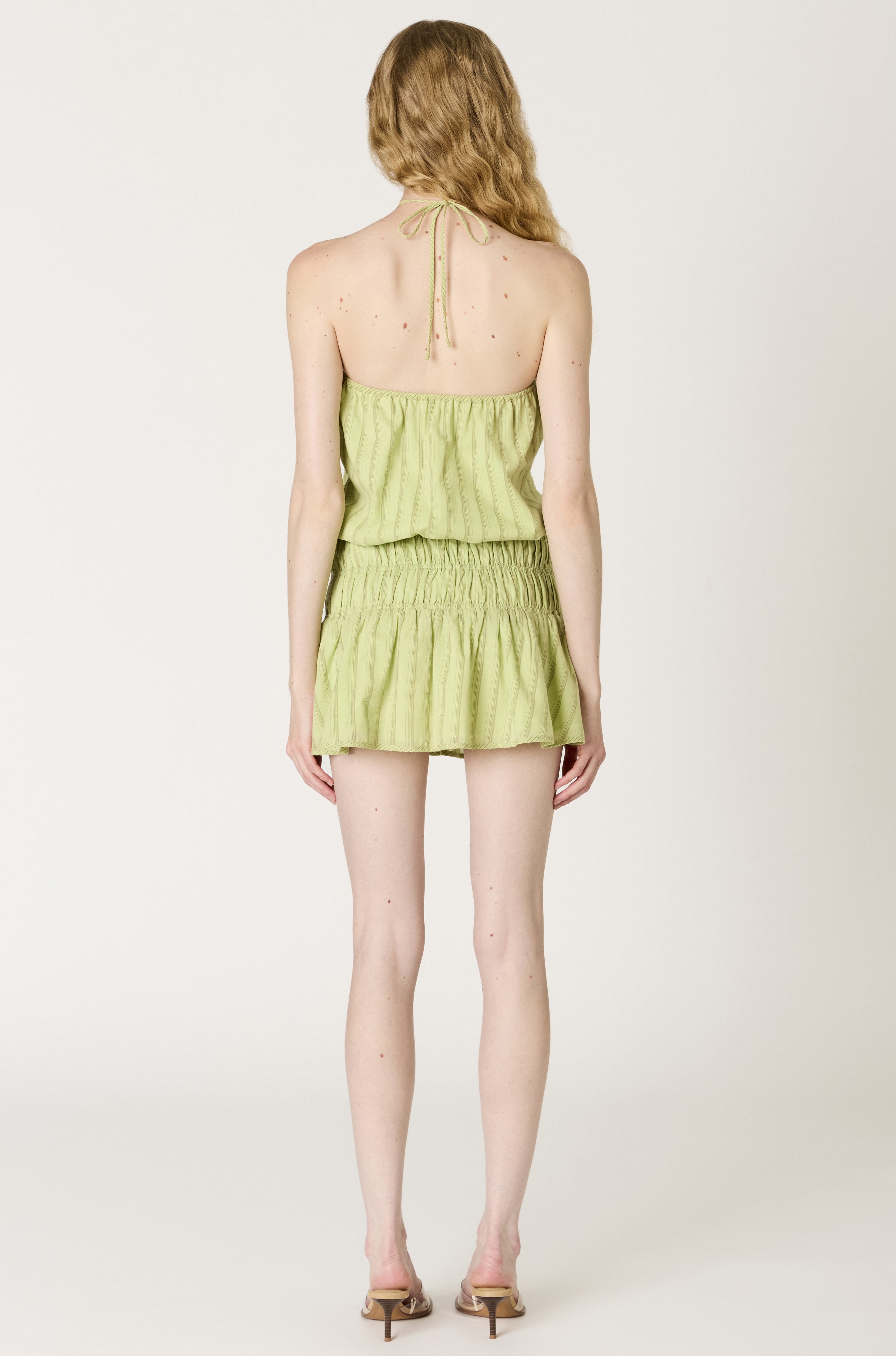 A woman with wavy blonde hair faces away, wearing the Nia Mara Dress Coverup—a light green cotton mini dress with a halter neck and open back—paired with beige heeled sandals against a plain white background.