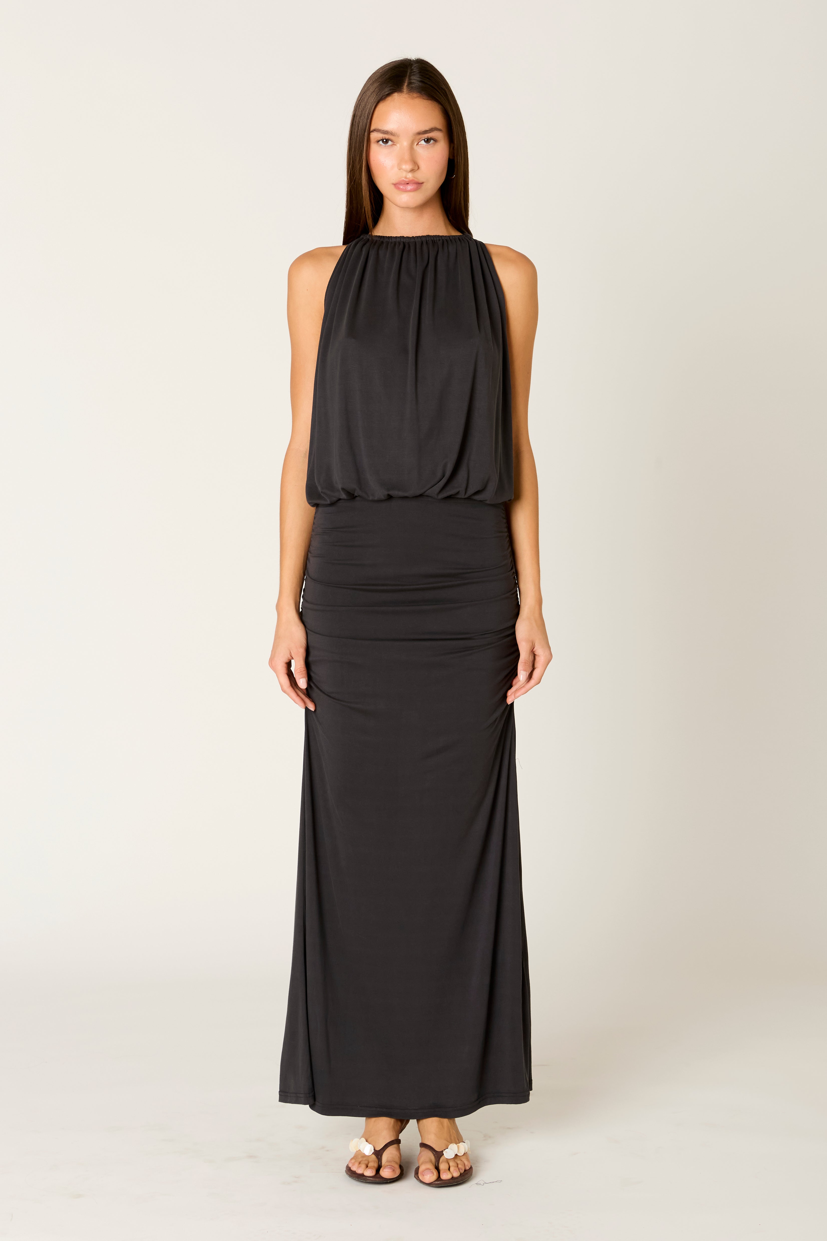 A woman stands facing forward, wearing the Nia Sebastiano Dress in ONYX—a sleeveless, floor-length black maxi dress with a blouson bodice paired with sandals—set against a plain light background.