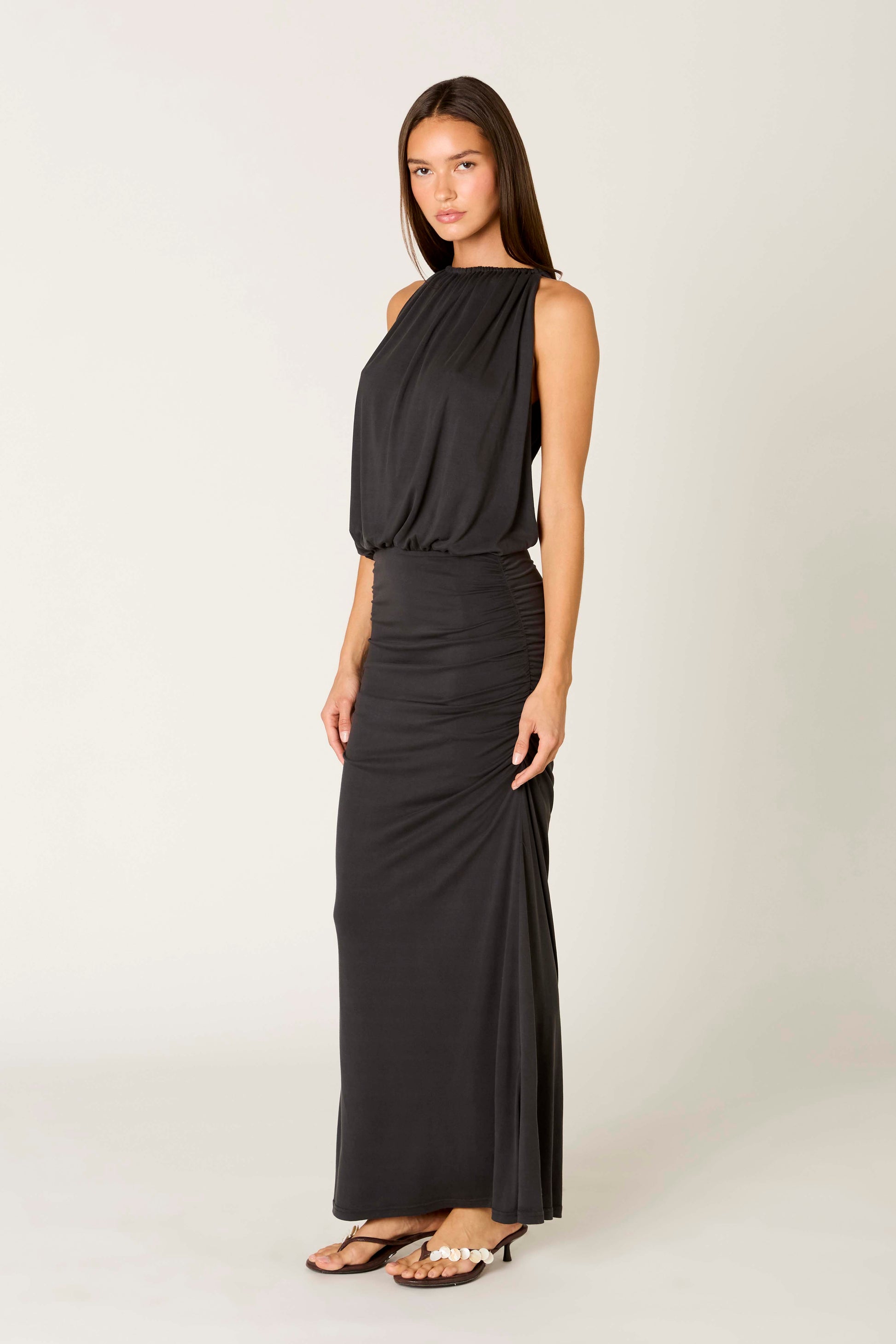 A woman stands against a plain light background wearing the Nia Sebastiano Dress—a sleeveless, floor-length black maxi style with a blouson bodice, gathered waist, and side ruching—paired with heeled sandals.