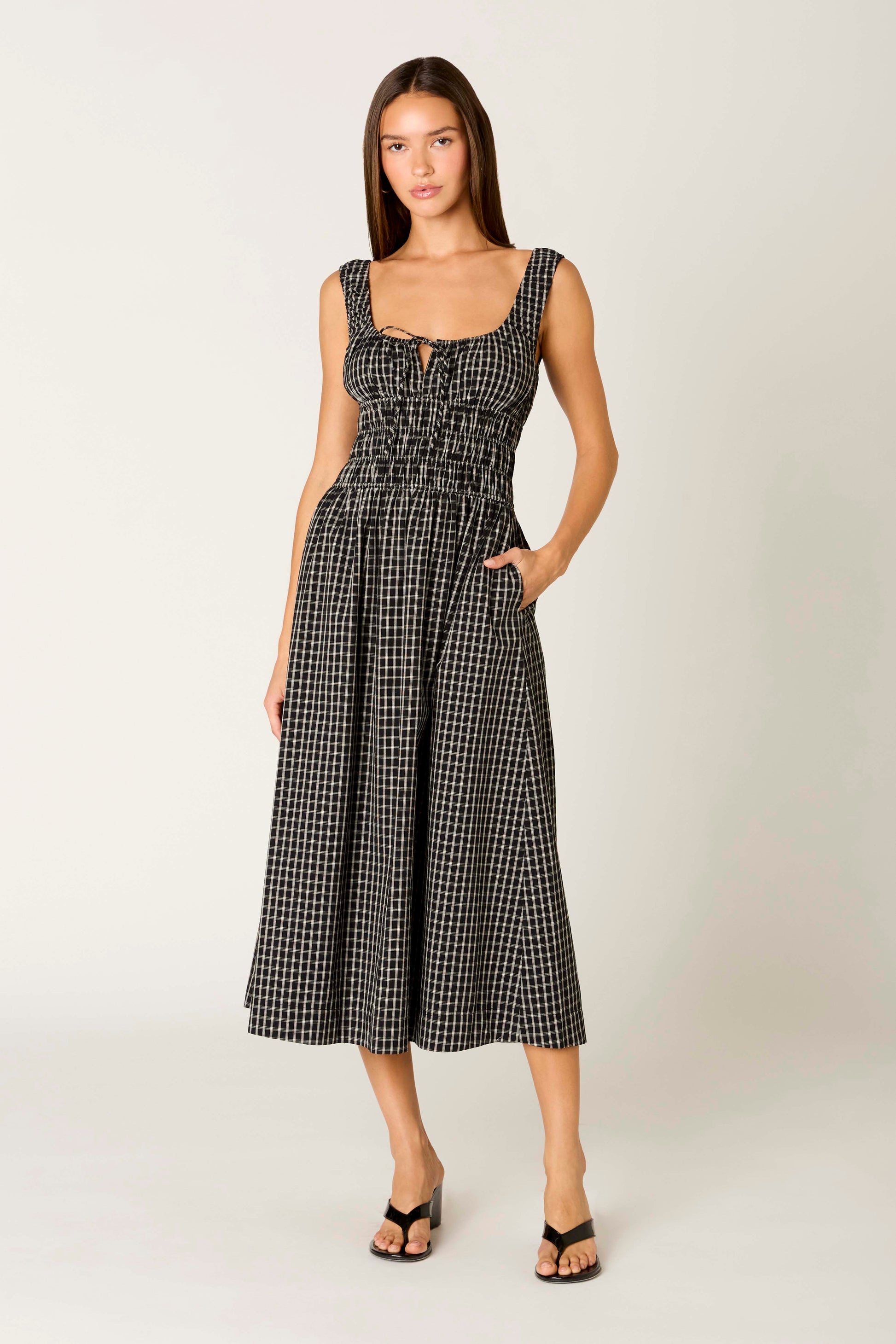 A woman with straight brown hair and a neutral expression wears the Salt Midi Dress by Nia in black, paired with black heeled sandals.