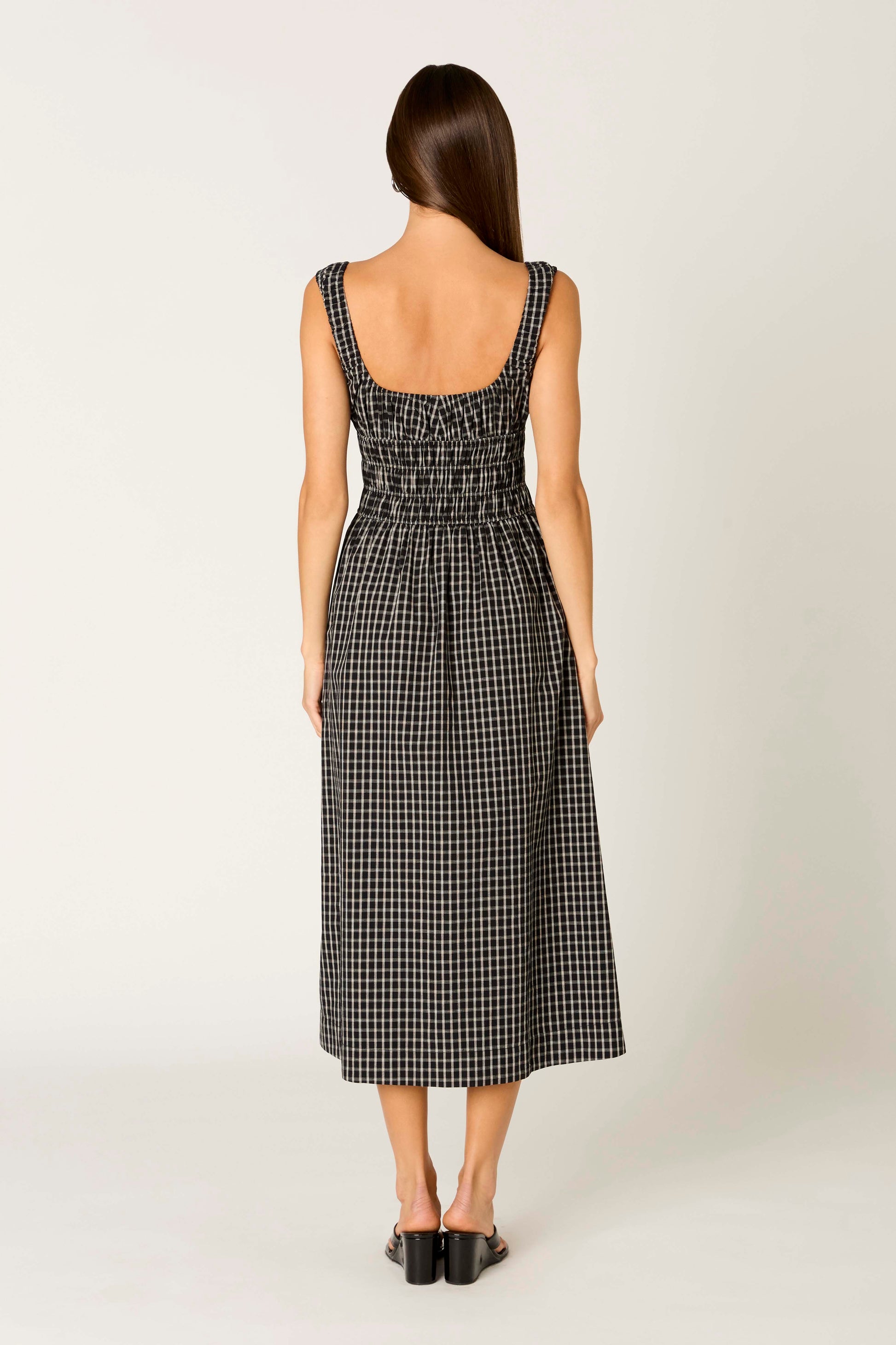 A woman faces away on a plain light background, wearing the Nia Salt Midi Dress—a sleeveless black and white gingham fit-and-flare with a smocked bodice—paired with black sandals.