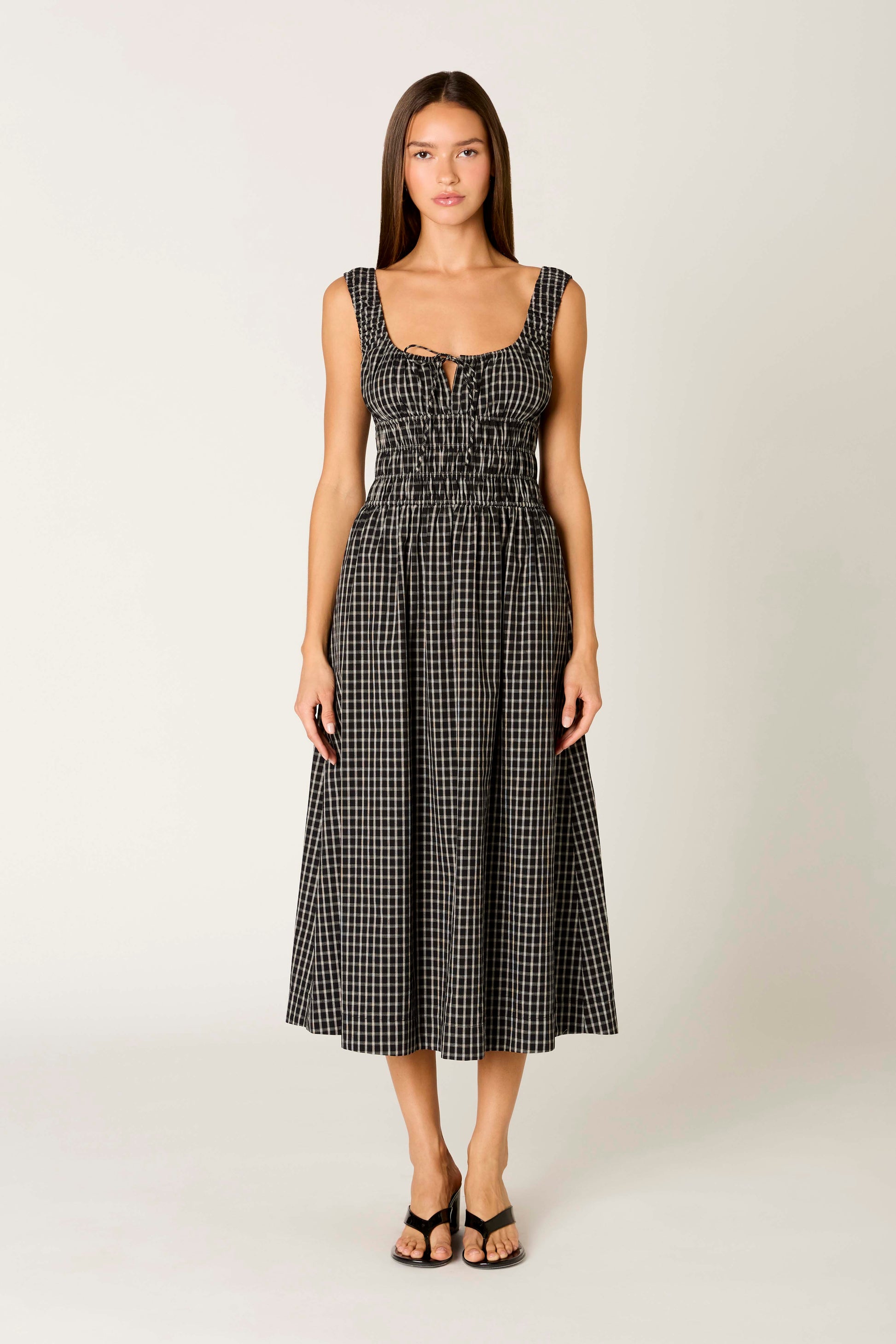 A woman faces forward against a light background, wearing the Nia Salt Midi Dress—a sleeveless black and white gingham fit-and-flare with gathered bodice and front cutout—paired with black open-toe heels.