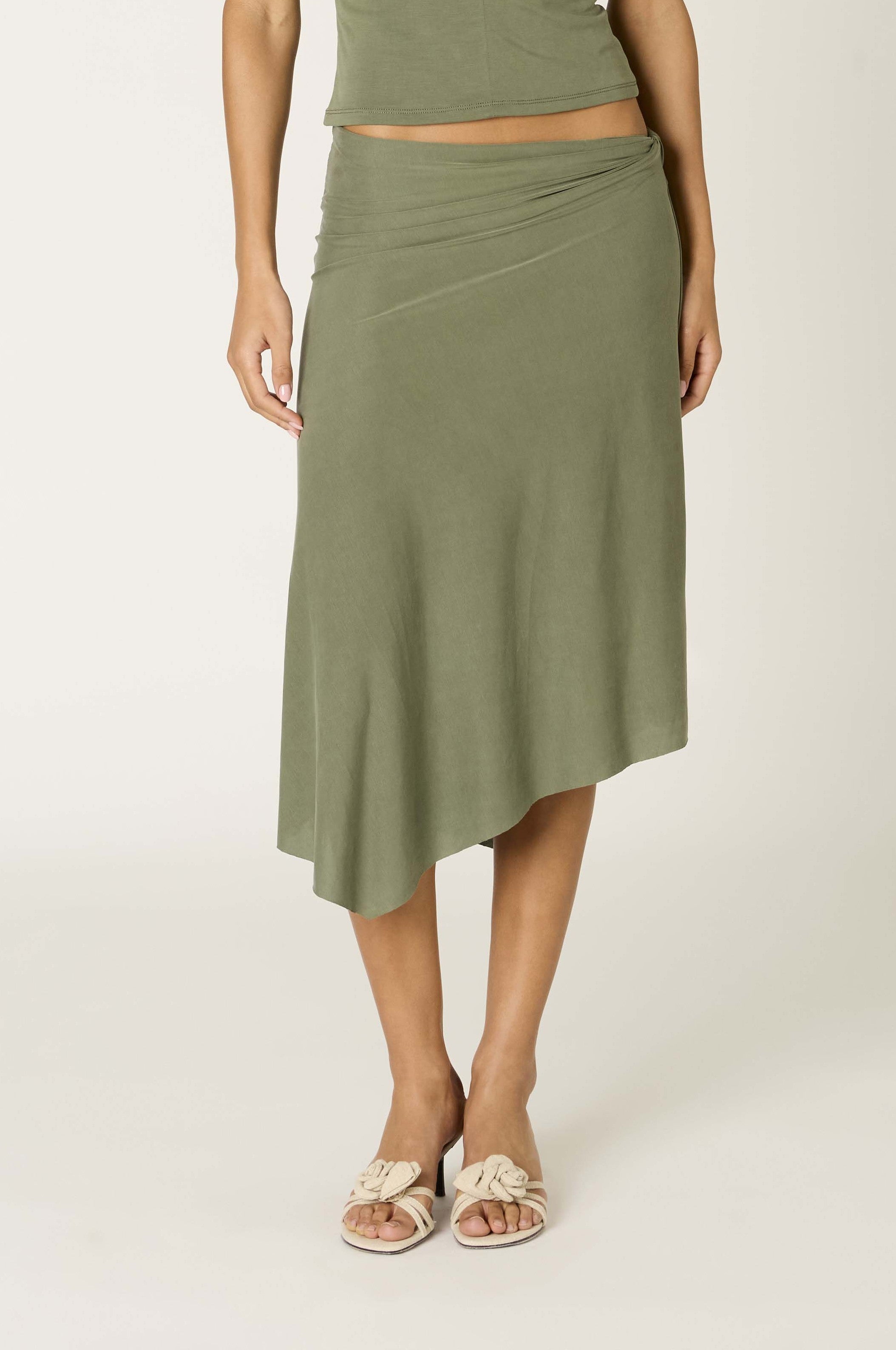 A person wearing the Nia Francesca Skirt in OLIVE, featuring an asymmetrical hem and a draped silhouette, paired with cream-colored sandals. The look is shown from torso to feet against a plain background, highlighting the modern elegance of the skirt’s design.