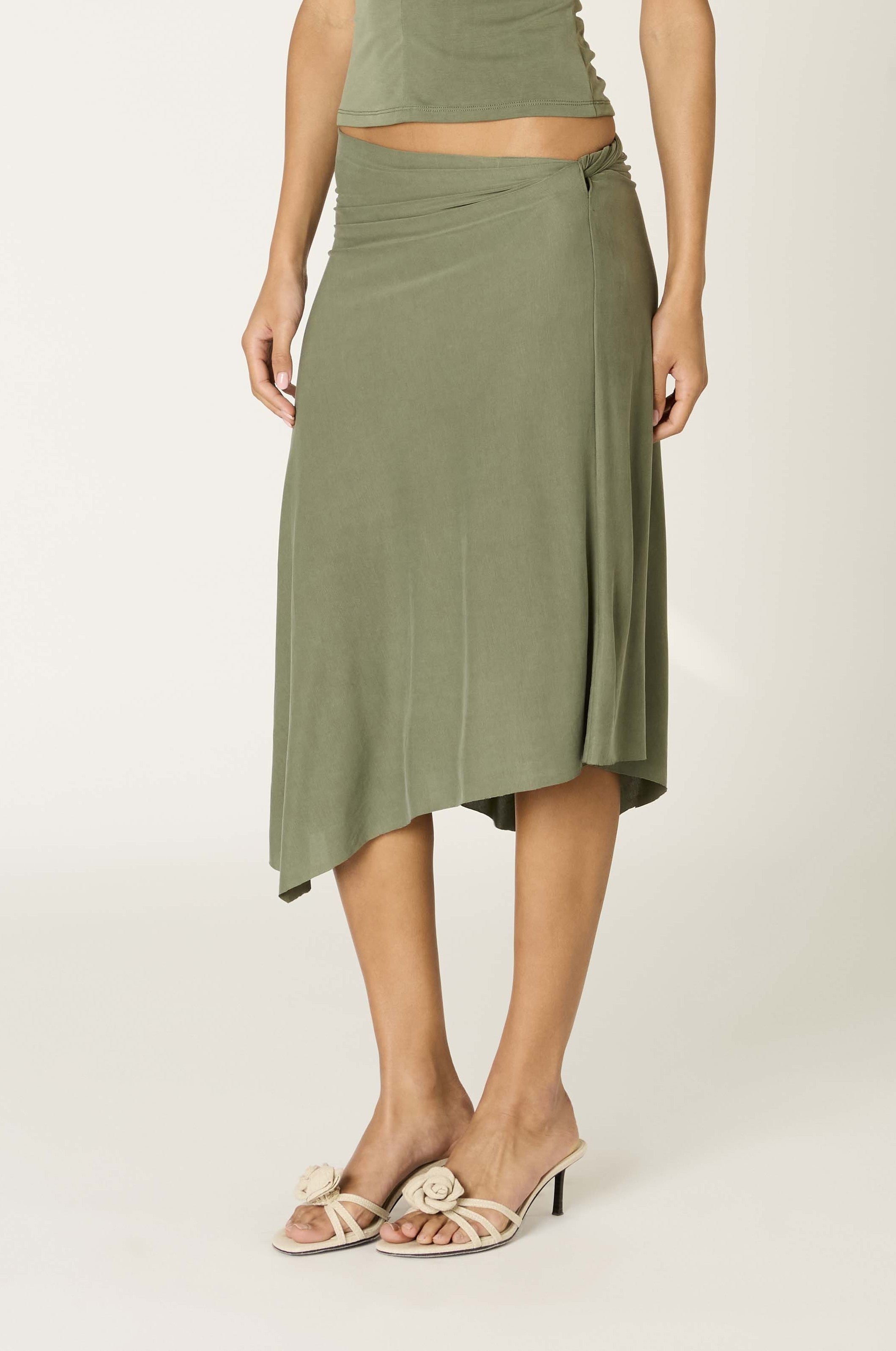 A woman is shown standing against a plain light background, wearing the Nia Francesca Skirt—an olive green midi skirt with an asymmetrical hem—and beige heeled sandals. Only her lower body is fully visible.
