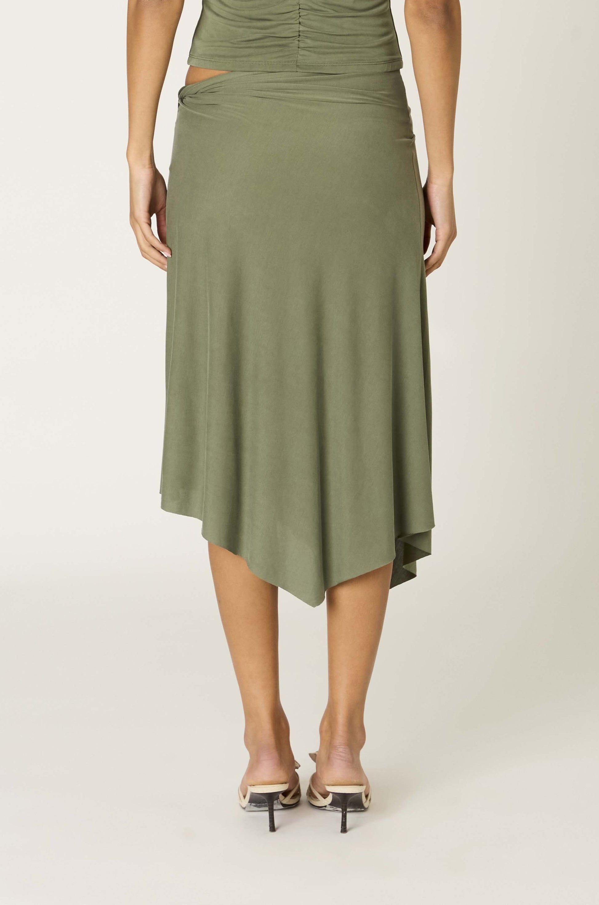 A person wearing the Nia Francesca Skirt—an olive green midi skirt with an asymmetrical hem—paired with heeled sandals, shown from the back against a plain light background.