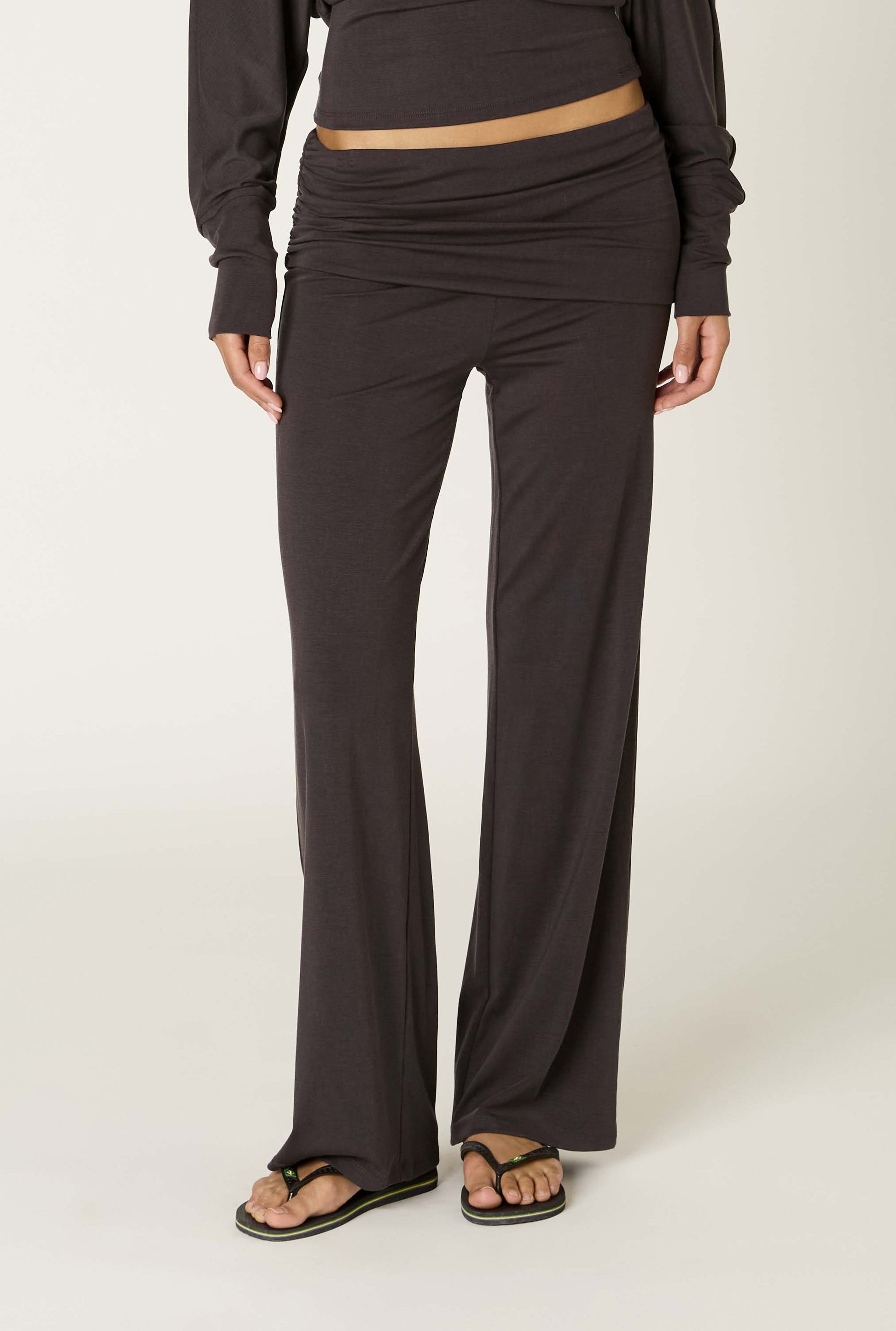 A person wearing the Delfina Pant by Nia in COAL—a high-waisted, relaxed-fit wide-leg pant made from soft Tencel fabric—paired with a matching long-sleeve top and black sandals, standing against a plain light background.