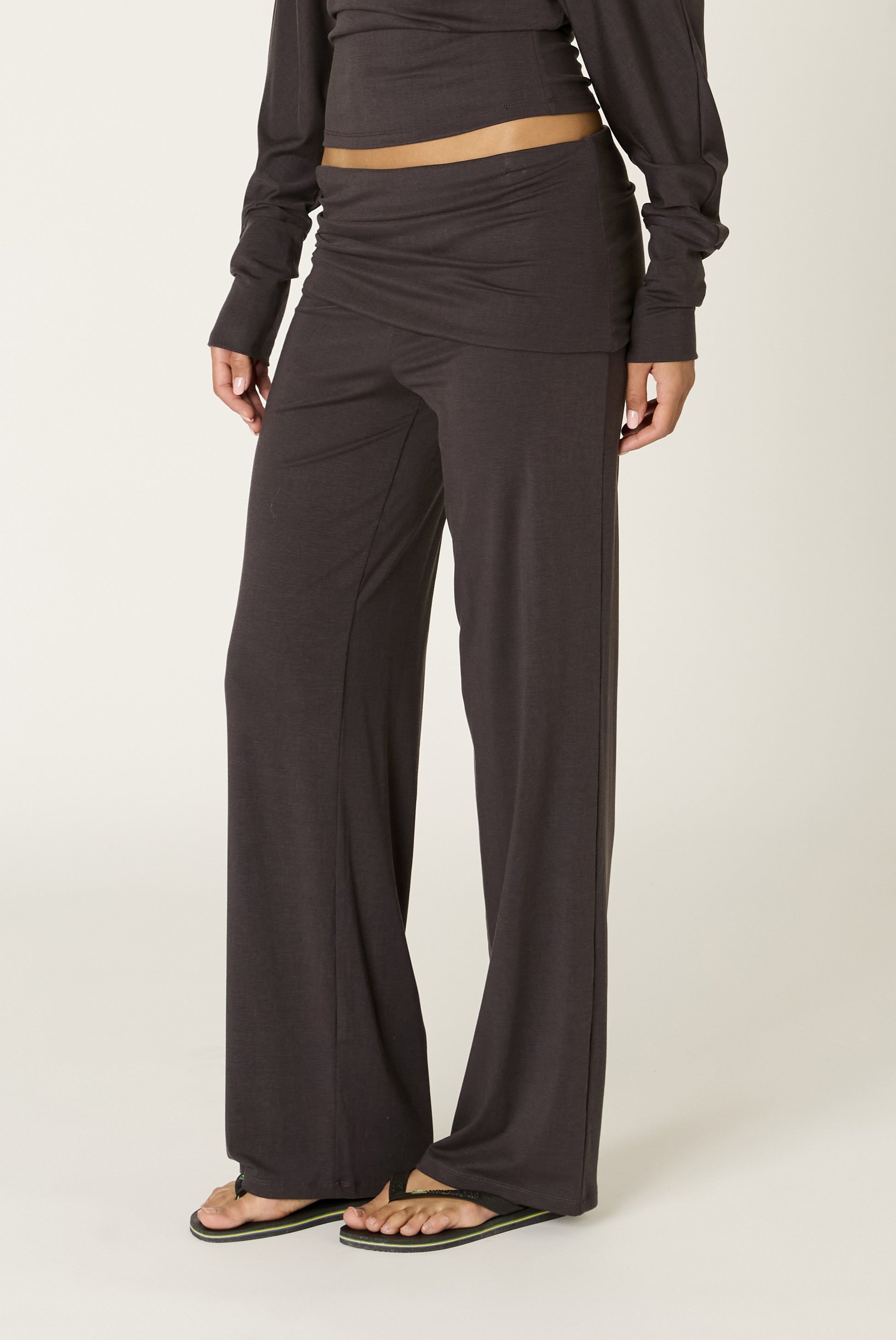 A person wearing the Nia Delfina Pant in a relaxed fit dark gray wide-leg style, paired with a matching long-sleeve crop top and black flip-flops, stands against a light background with only their lower half visible.