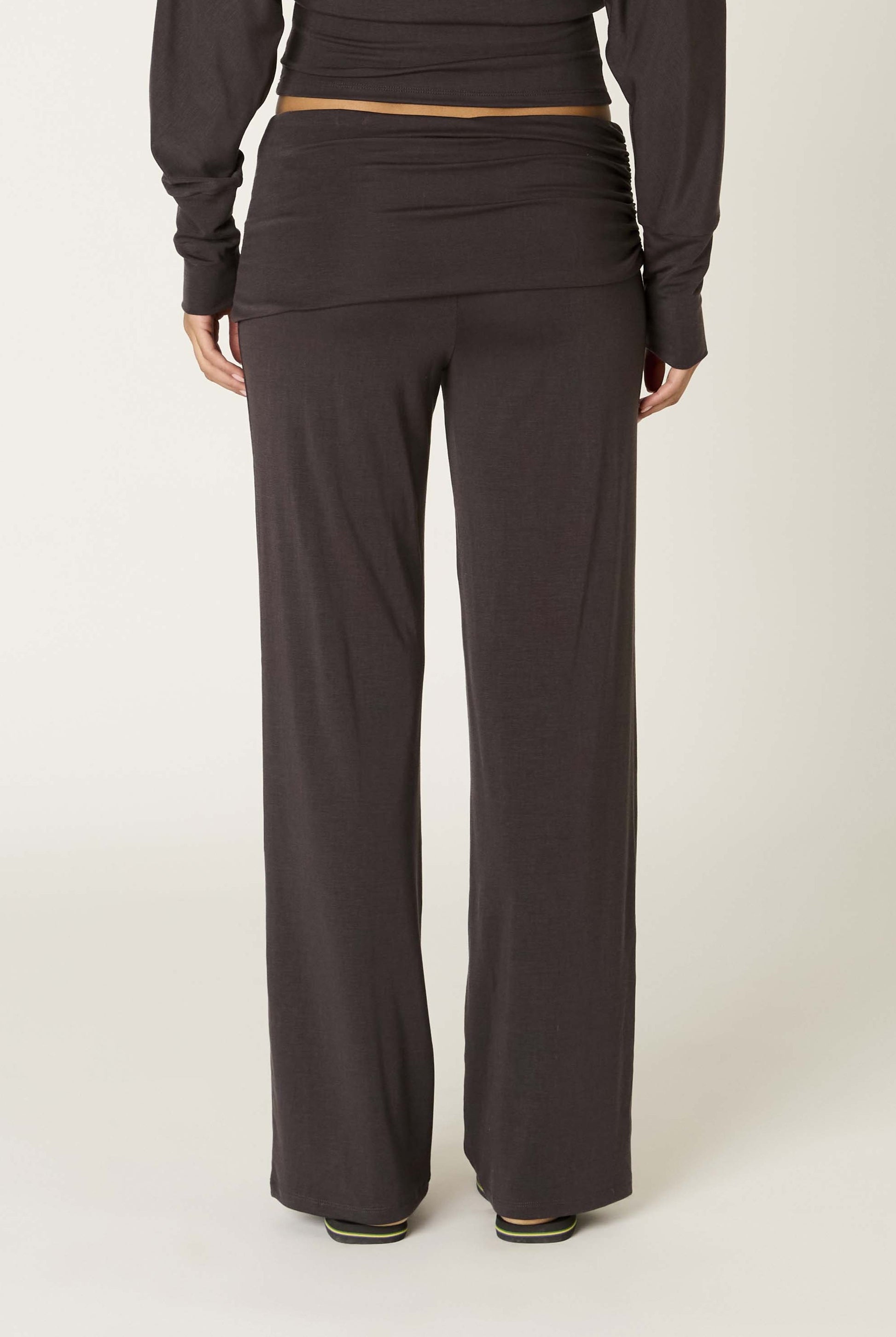 A person is shown from the back, standing against a plain light background, wearing Nia's Delfina Pant in dark brown Tencel with a relaxed fit and a coordinating long-sleeve top.