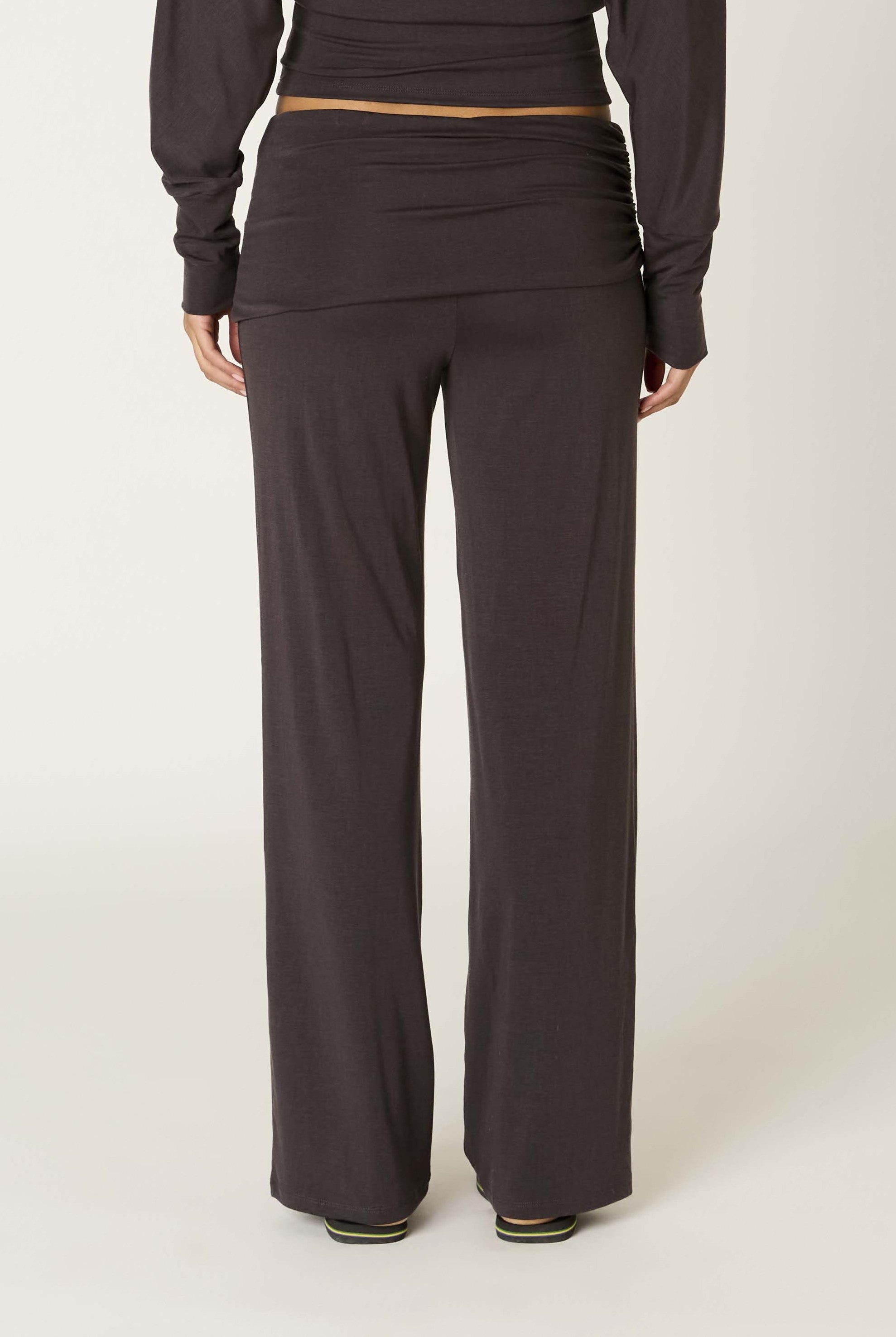 A person is shown from the back, standing against a plain light background, wearing Nia's Delfina Pant in dark brown Tencel with a relaxed fit and a coordinating long-sleeve top.