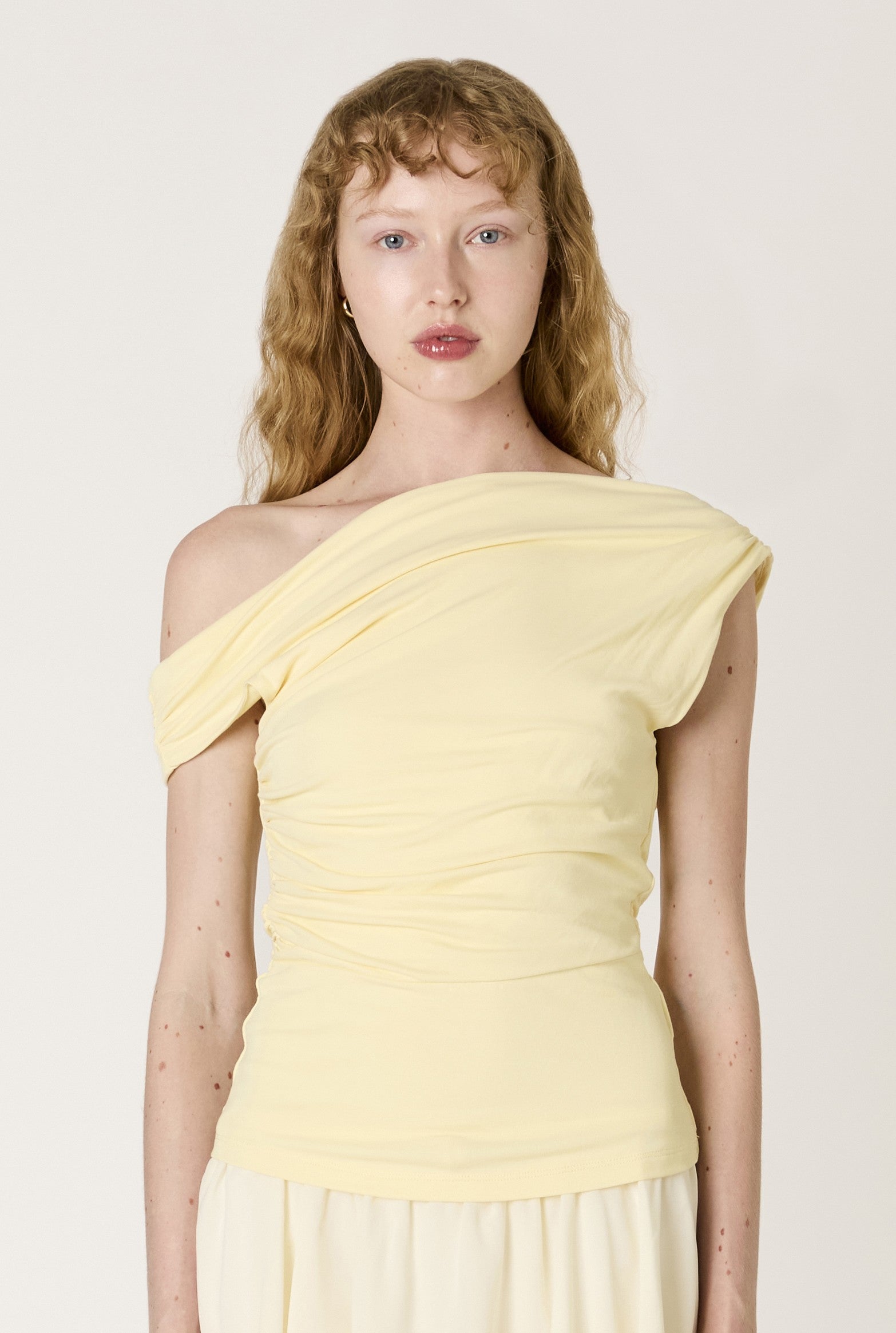 A person with long, wavy blonde hair wears the Nia Mariposa Top, a pale yellow one-shoulder stretch piece, standing against a plain light background and looking directly at the camera with a neutral expression.