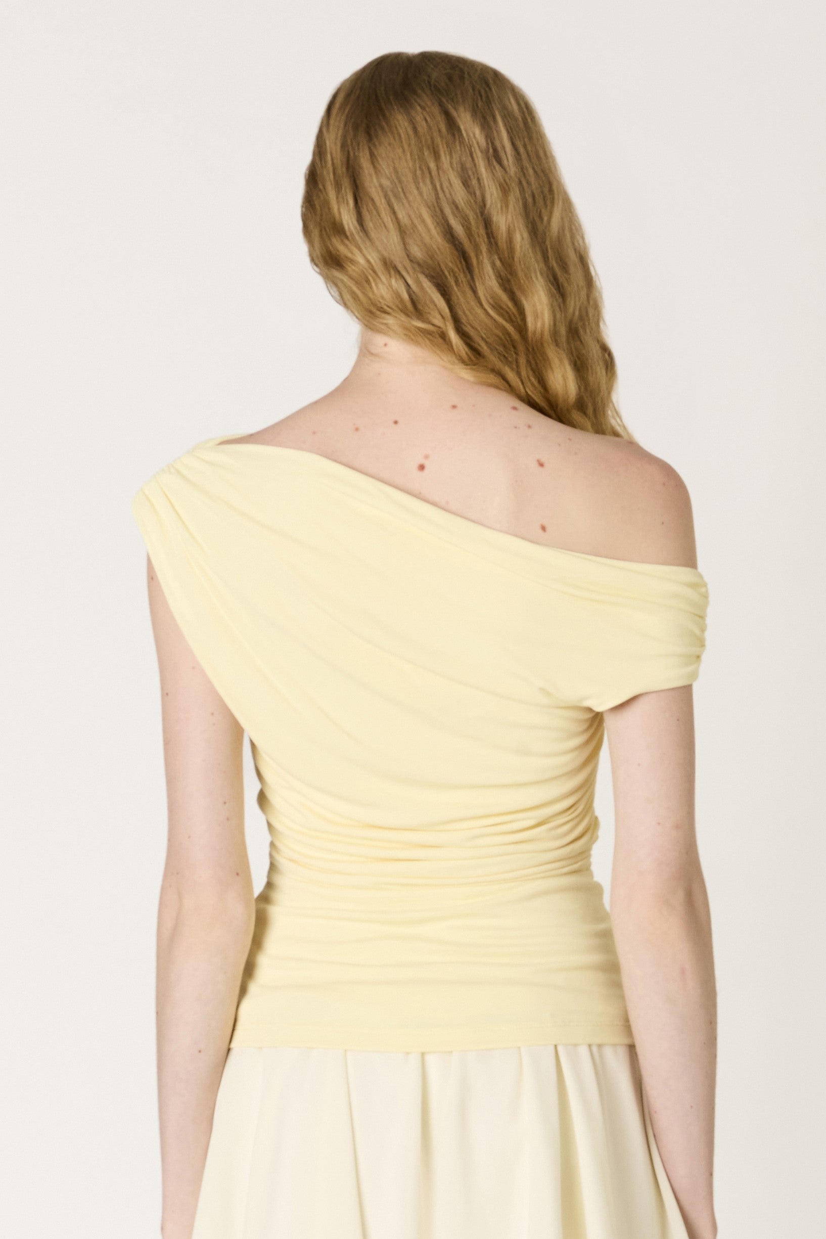 A woman with wavy blonde hair is seen from behind, wearing the pale yellow, body-hugging Mariposa Top by Nia. The background is plain and light-colored.