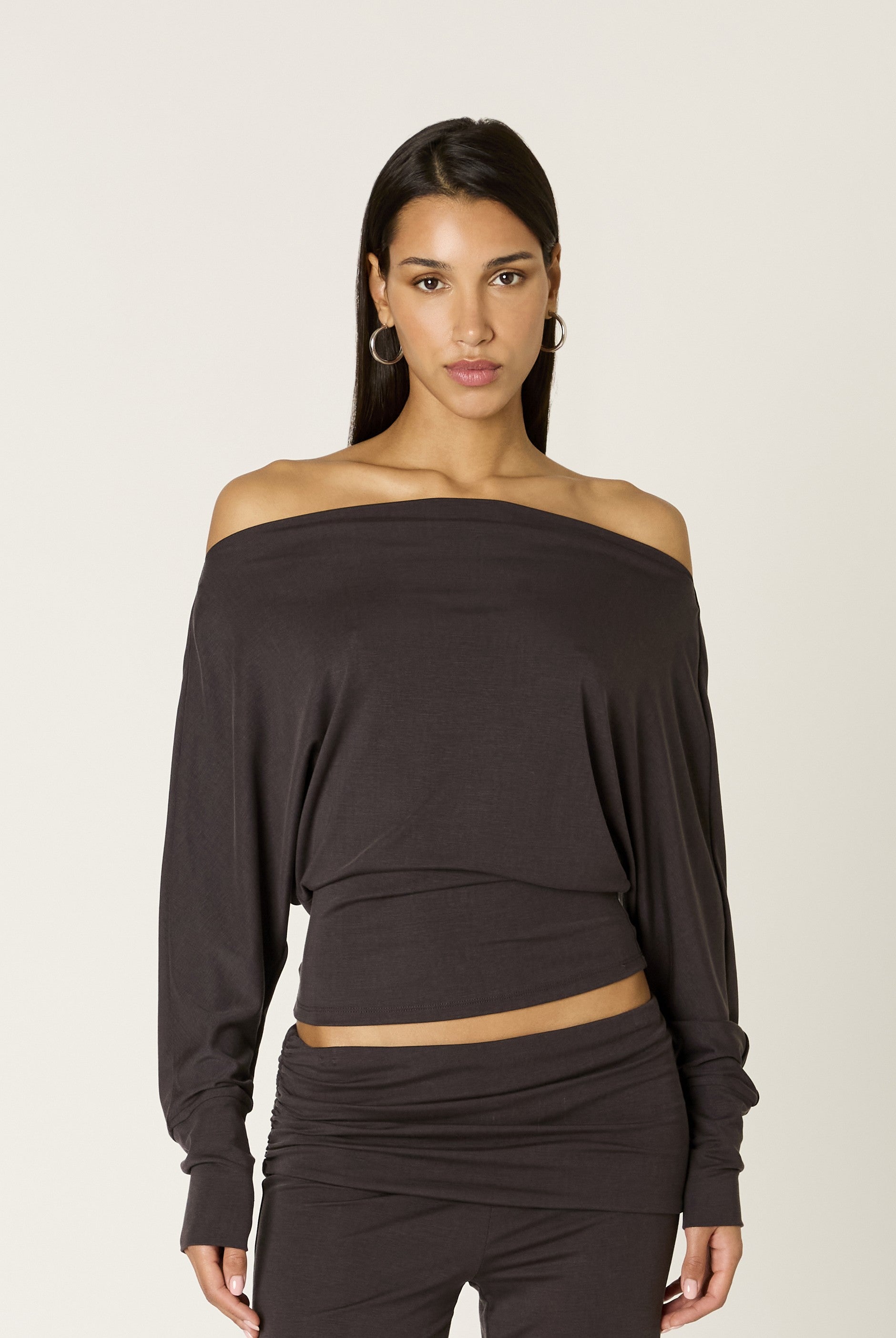 A woman wearing the Nia Elisabetta Top in COAL, featuring a dark off-the-shoulder neckline and long sleeves, paired with matching pants in a subtle blouson silhouette, stands against a plain light background, facing forward with a neutral expression.