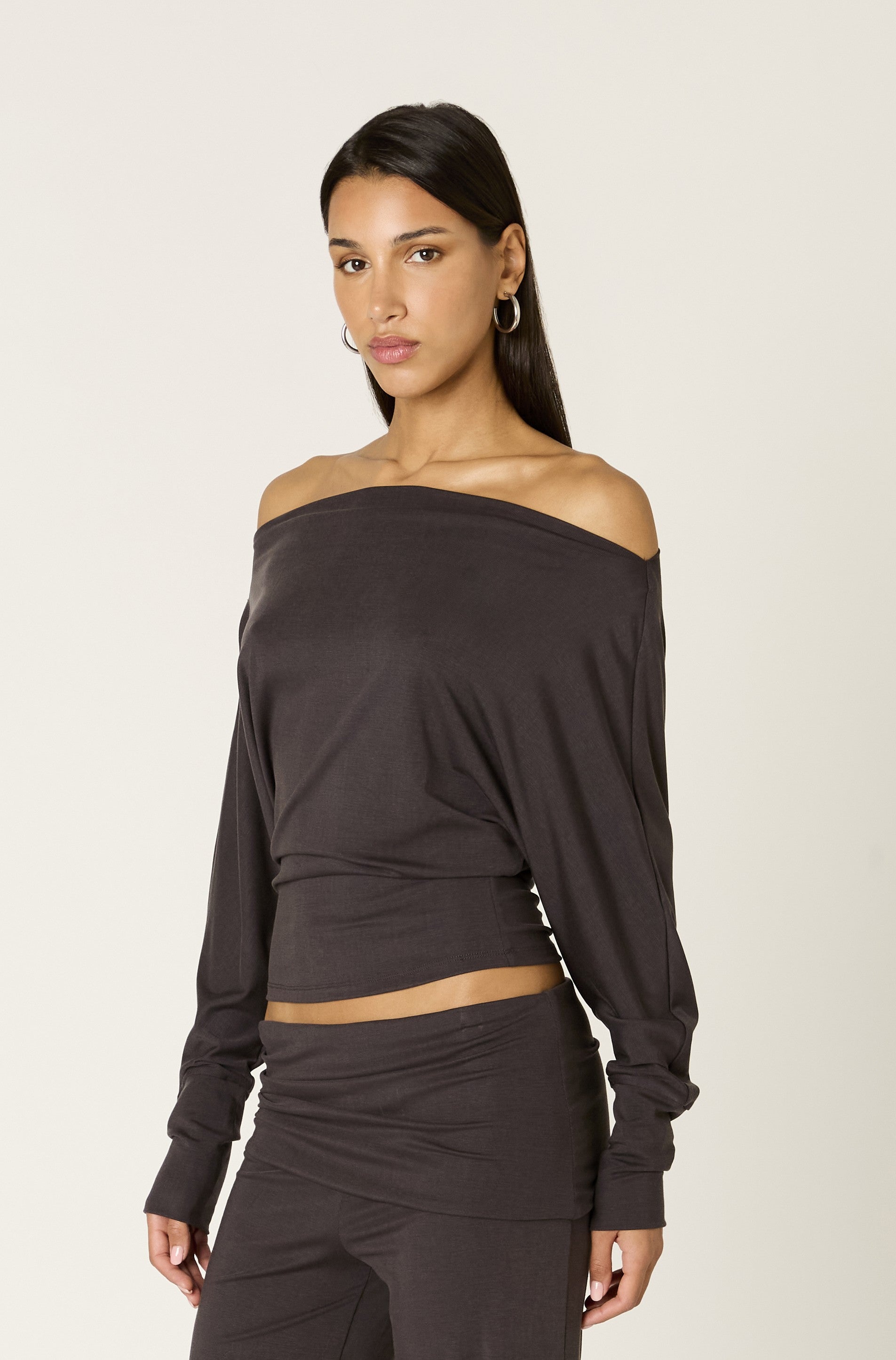A woman with long dark hair, wearing the Nia Elisabetta Top with an off-the-shoulder neckline and matching pants, stands against a plain light background, gazing into the camera with a neutral expression.