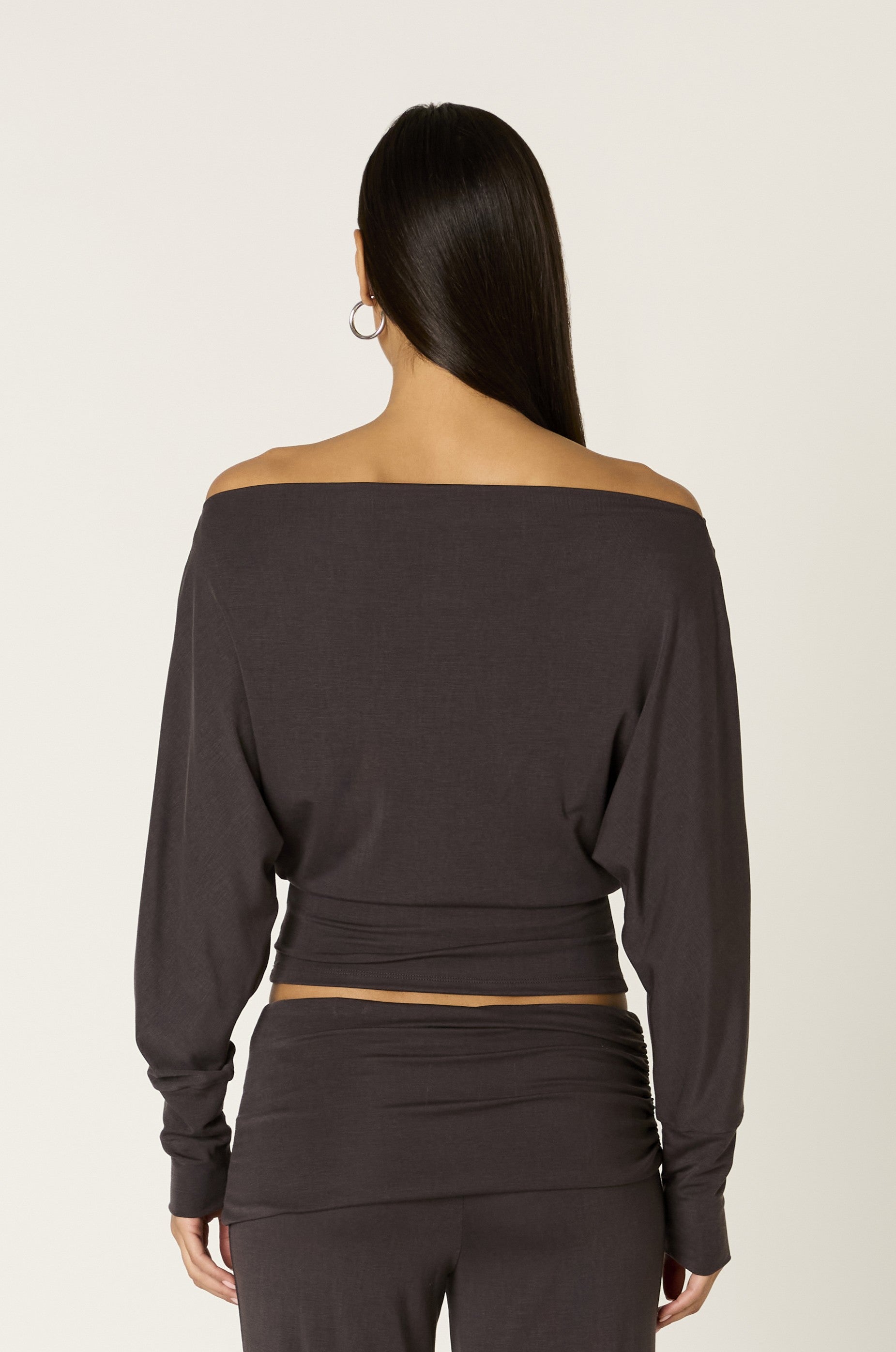A woman with long dark hair stands facing away, wearing the Nia Elisabetta Top—a Tencel, off-the-shoulder long sleeve top—paired with matching pants. She has hoop earrings and her arms are relaxed at her sides.